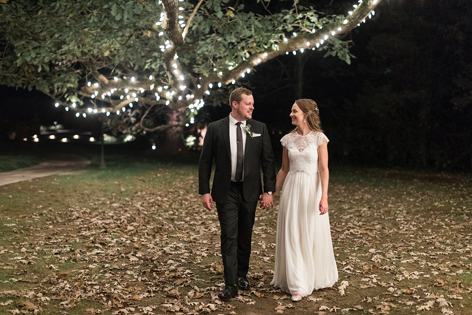 newlyweds hold hands walking on lawn at night at Aldie Mansion 