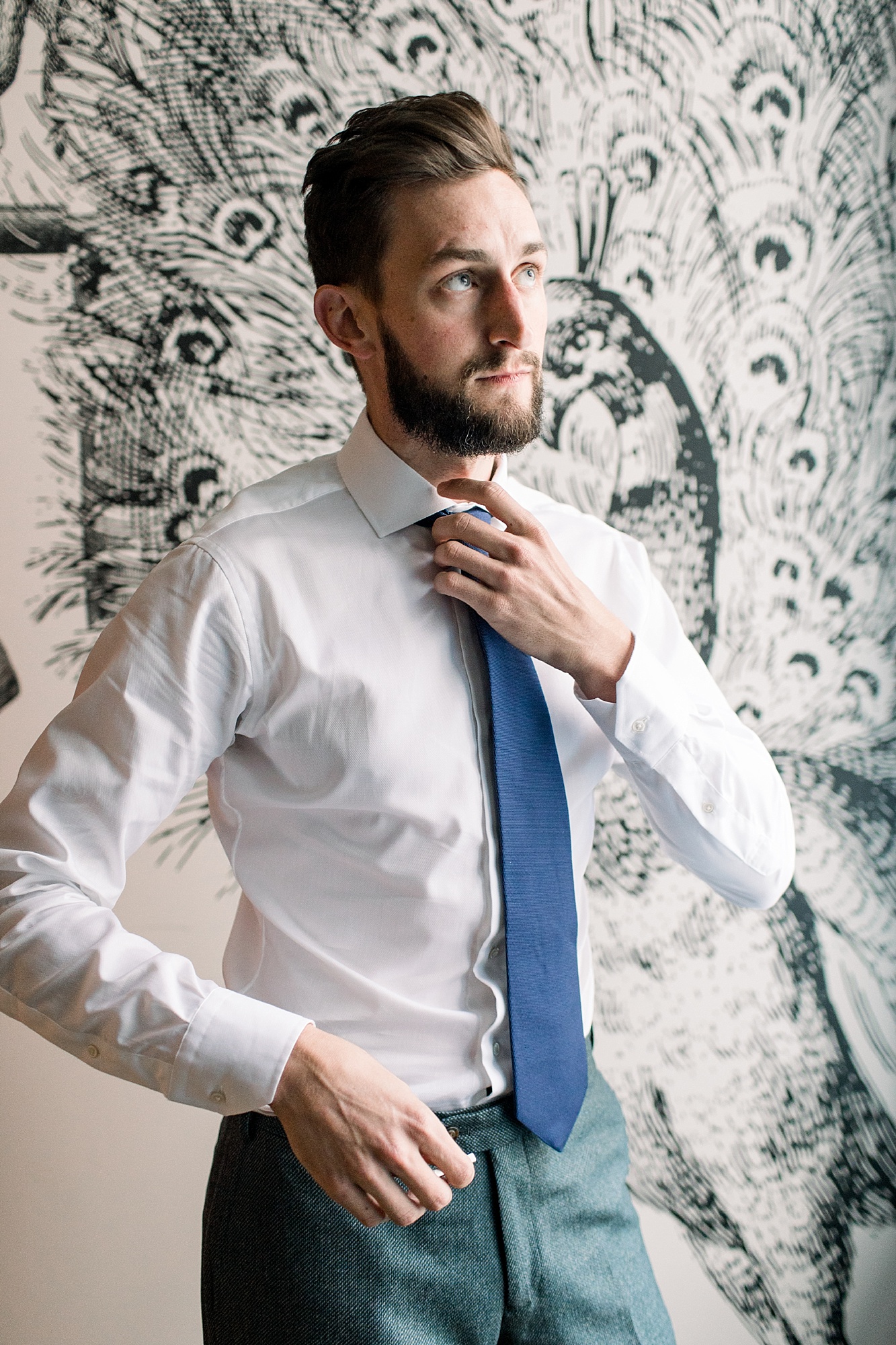groom adjusts blue tie in front of black and white wallpaper at The Hotel Monaco