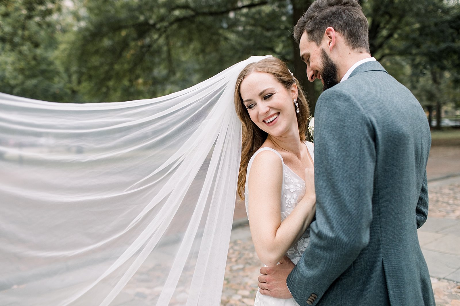 groom in grey suit jacket smiles at bride with veil floating behind them 