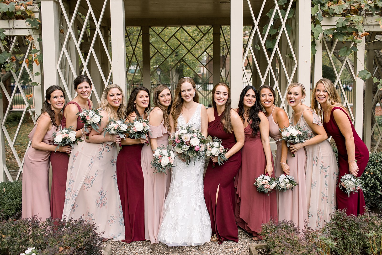 bride poses with bridesmaids in mismatched burgundy and pink gowns by gazebo