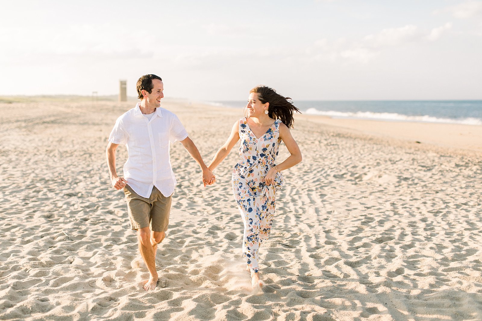 woman in white floral dress holds man's hand running down beach 