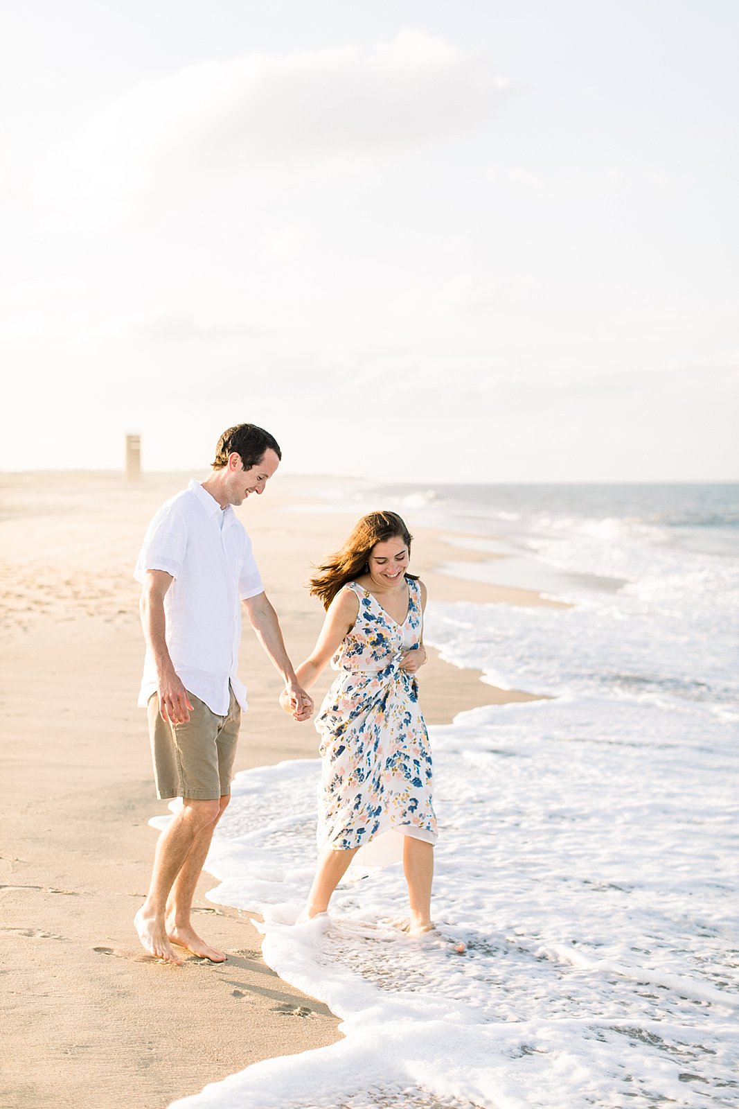 woman and man hold hands walking on Rehoboth Beach in waves during engagement session 