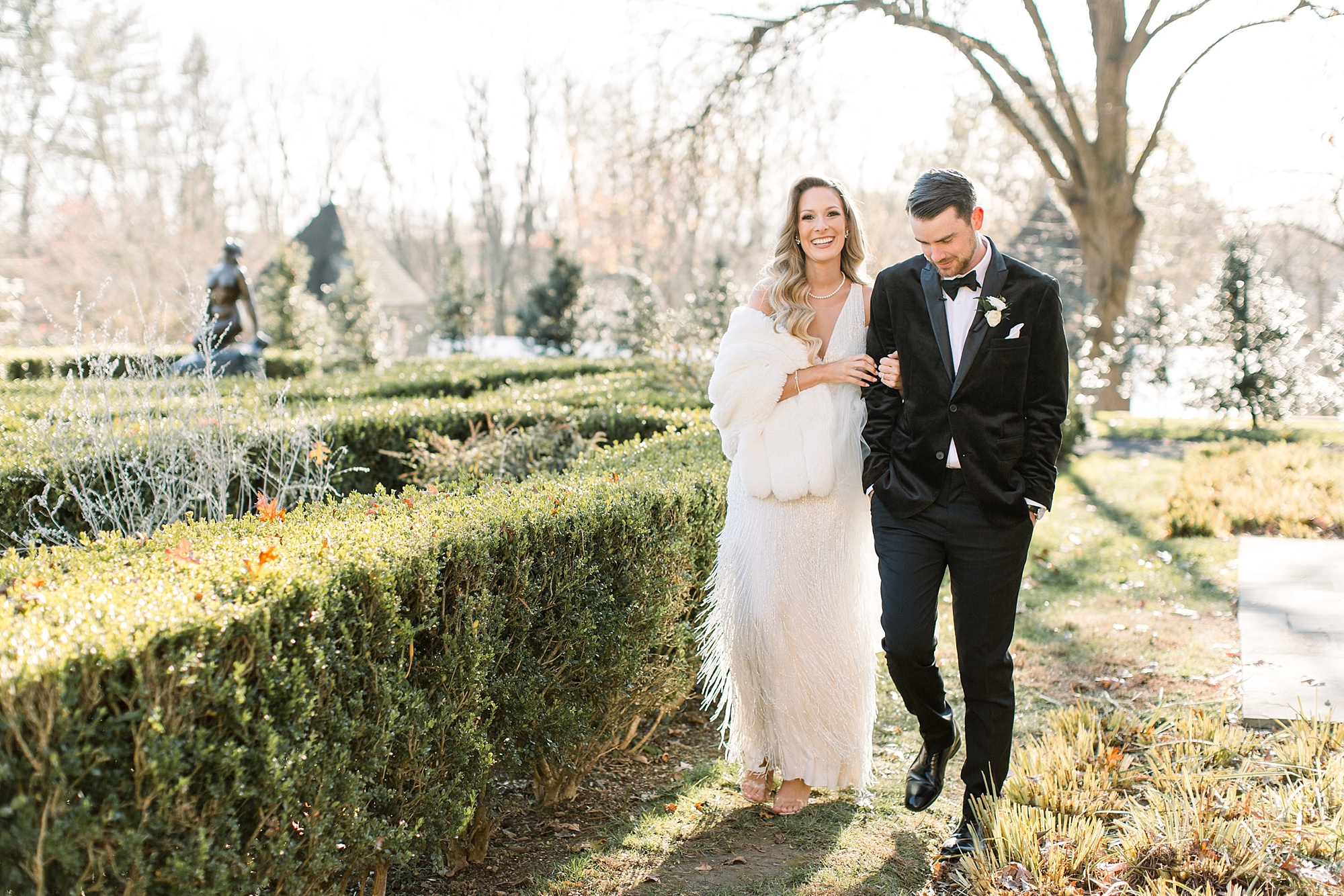 bride in fringe wedding dress with fur wrap walks with groom in black velvet suit at Tyler Gardens 
