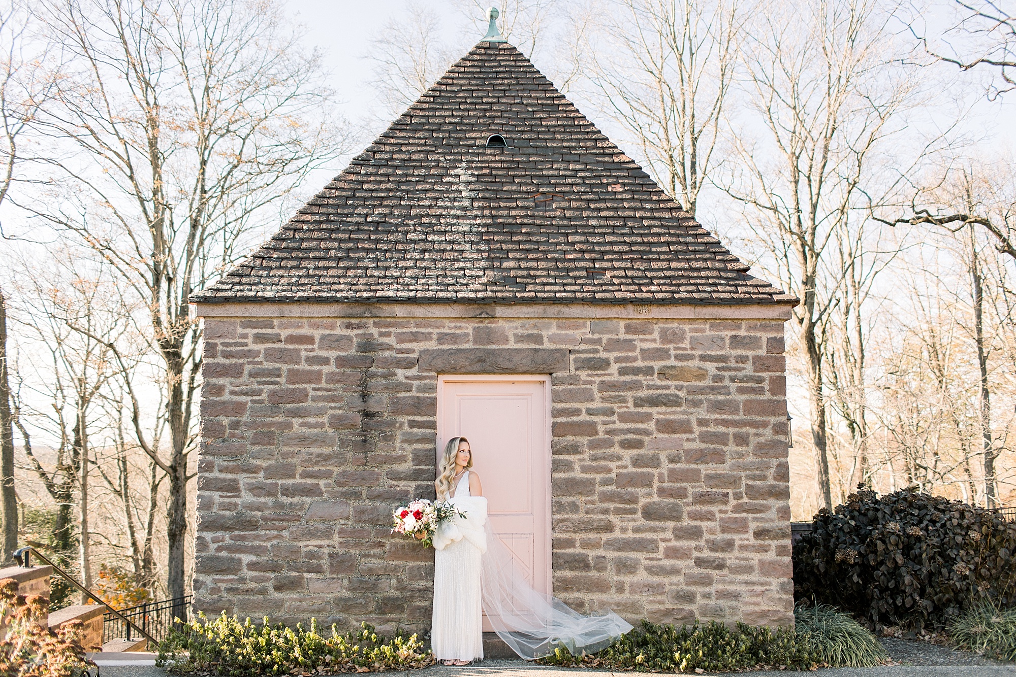 bride in fringe wedding dress with white wrap holds bouquet of pink and red flowers stands by pink door in stone wall 