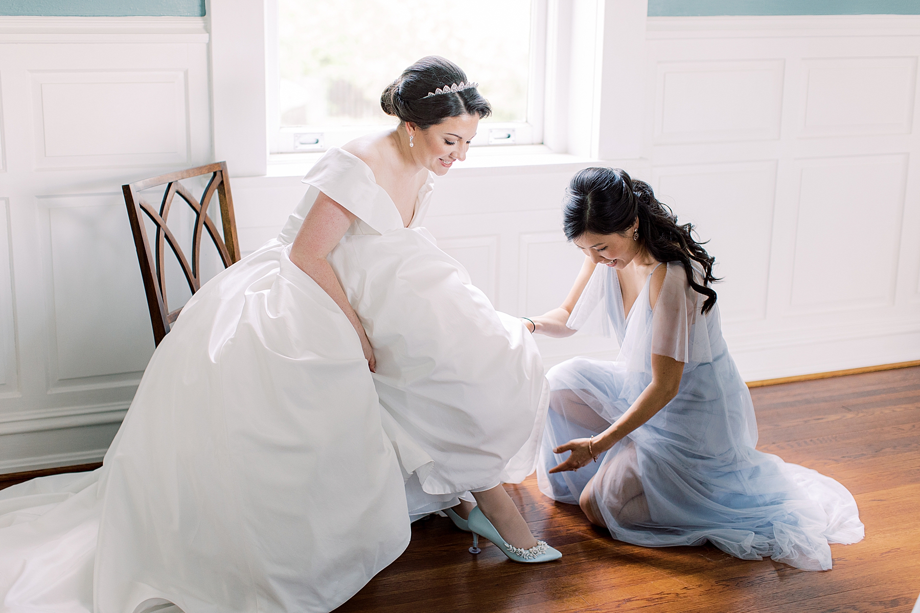 bridesmaid in dusty blue gown helps bride into blue shoes on wedding day 