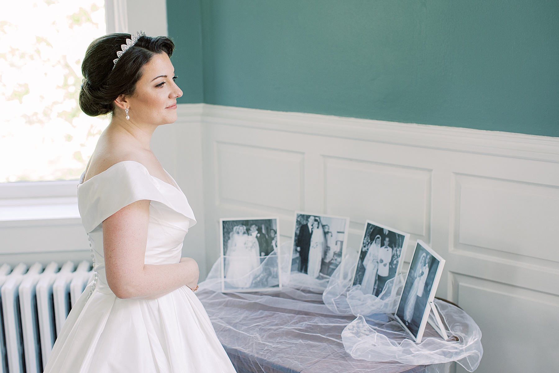 bride in off-the-shoulder gown stands by portraits of parents and grandparents on wedding day 