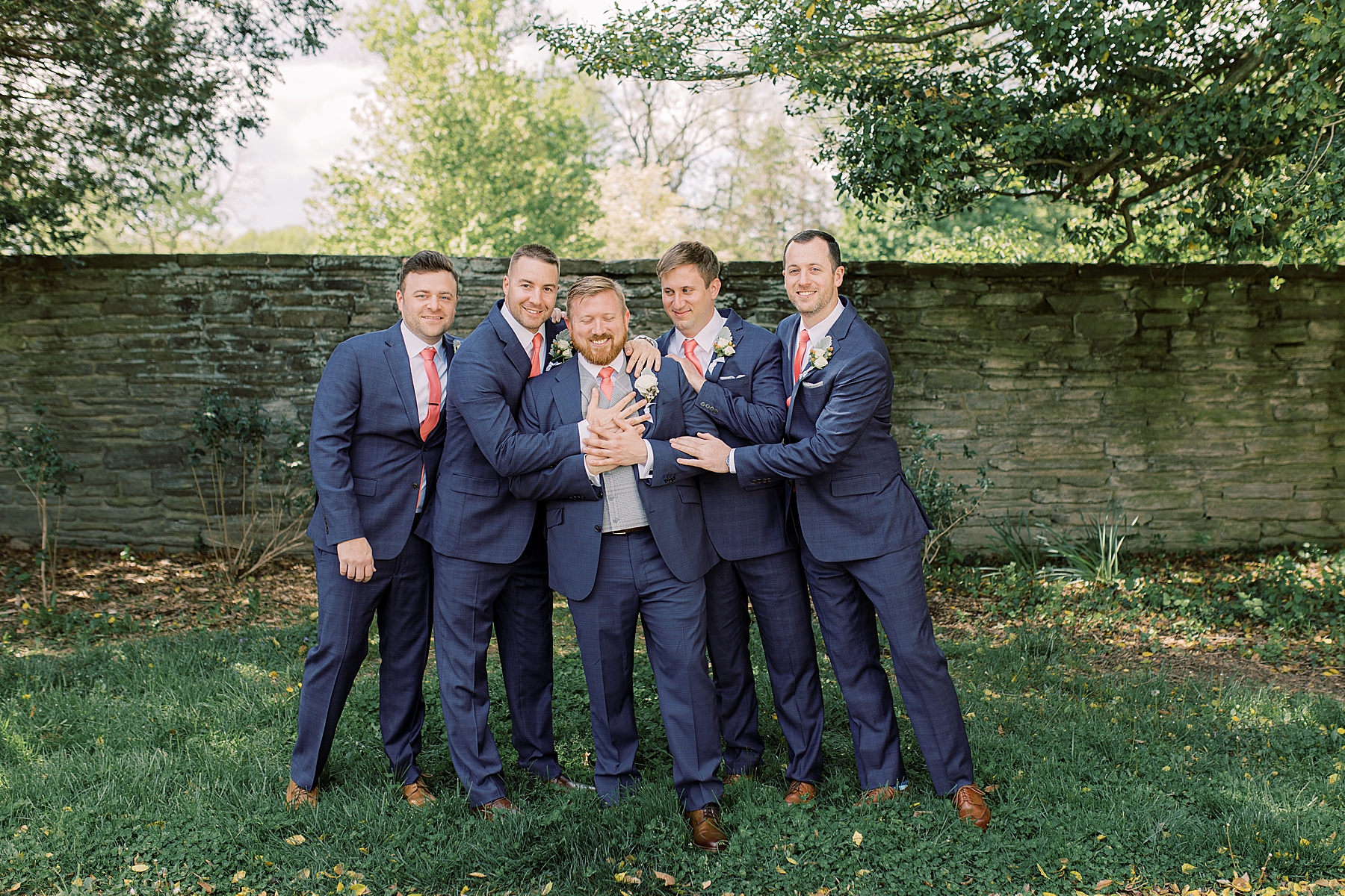 groomsmen in blue suits laugh with groom in front of stone wall at Bellevue Hall