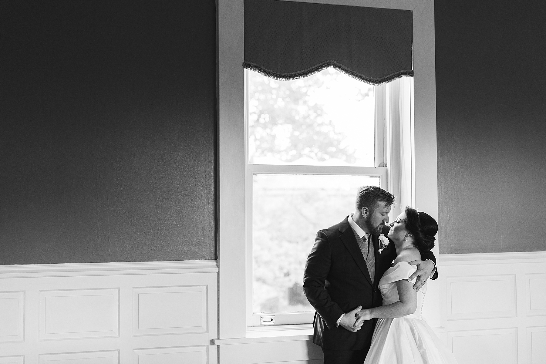 newlyweds hug by window leaning in for a kiss 
