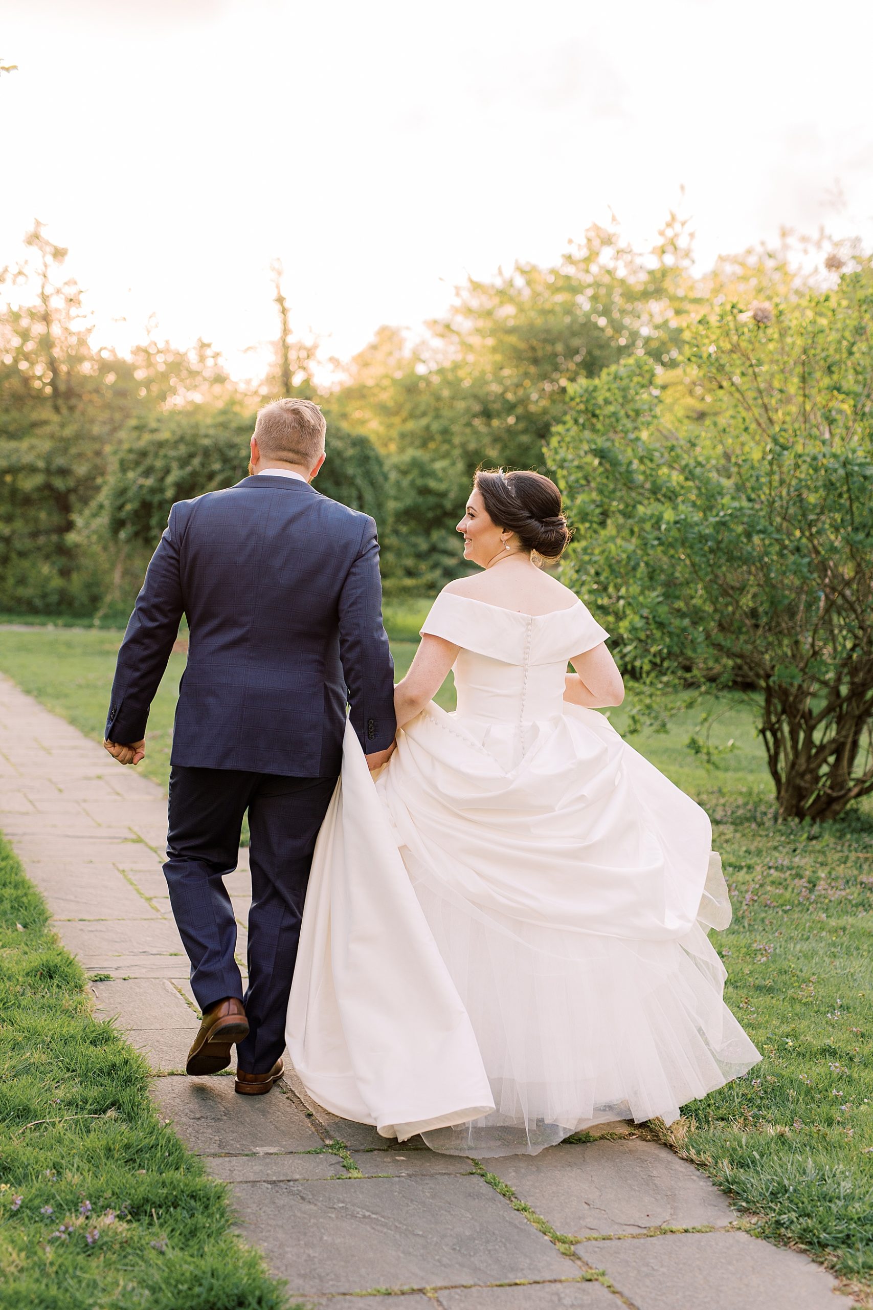 couple walks on brick path while bride carries skirt of wedding dress