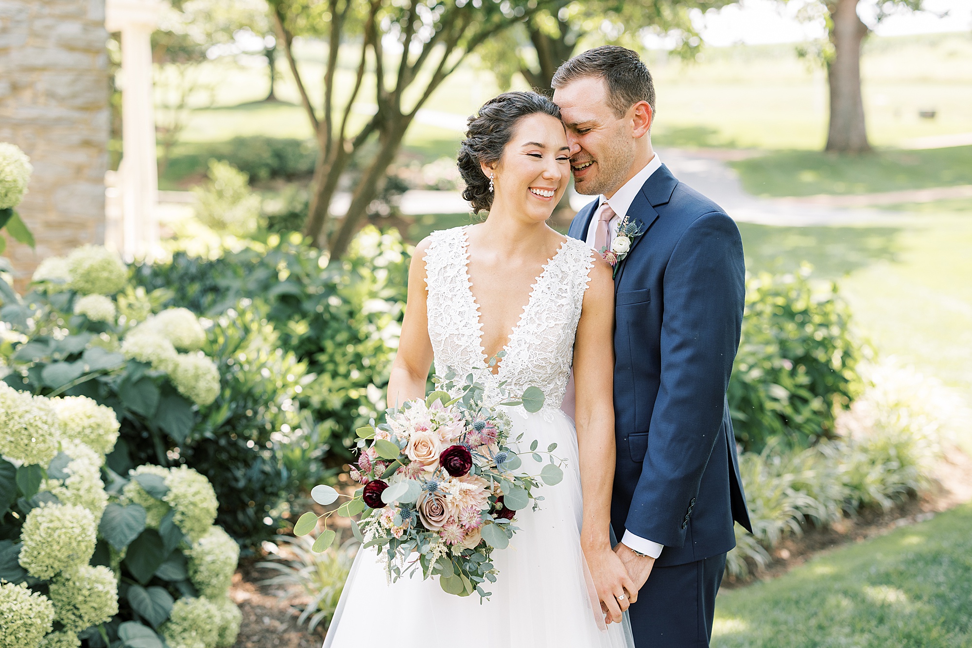 groom holds bride's hand and nuzzles her cheek by hydrangeas 
