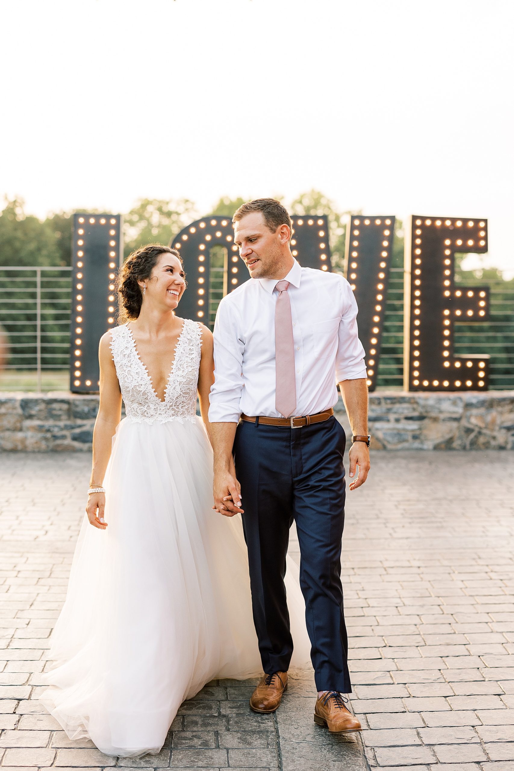 newlyweds hold hands walking in front of lit LOVE sign 