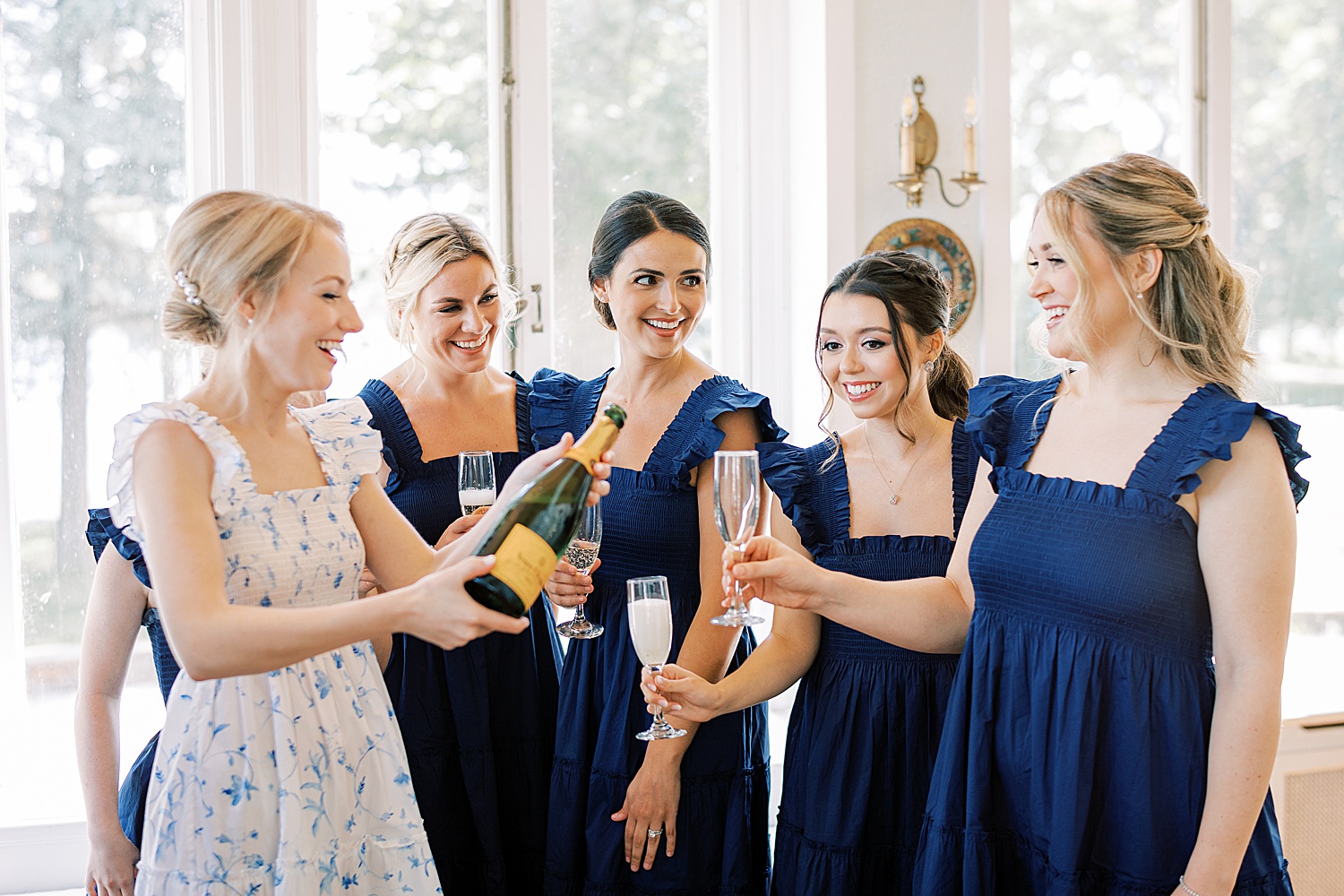 bride in blue and white dress pours champagne for bridesmaids in blue dresses 