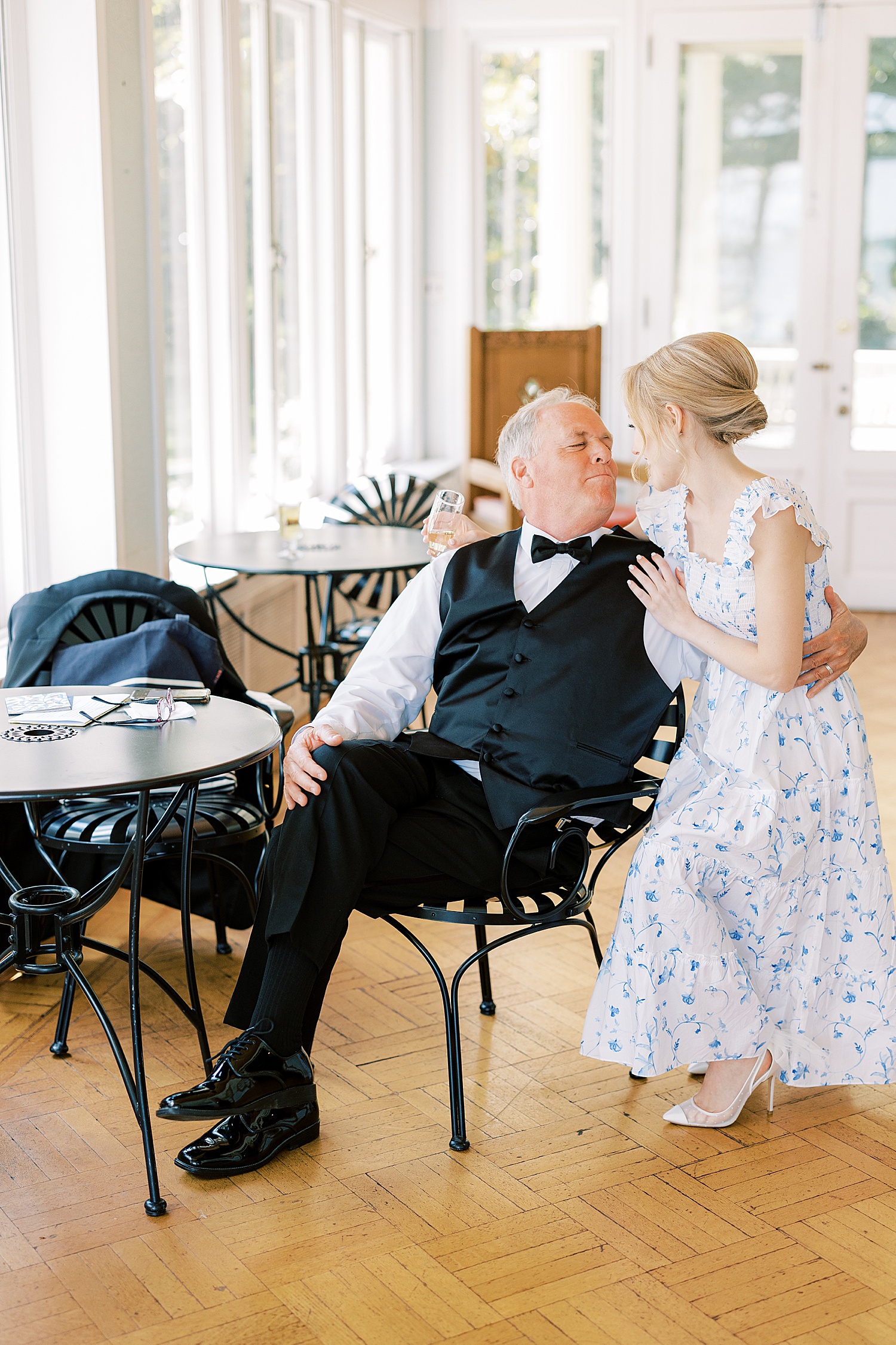 bride kneels down to talk to father in black chair inside room with big windows 