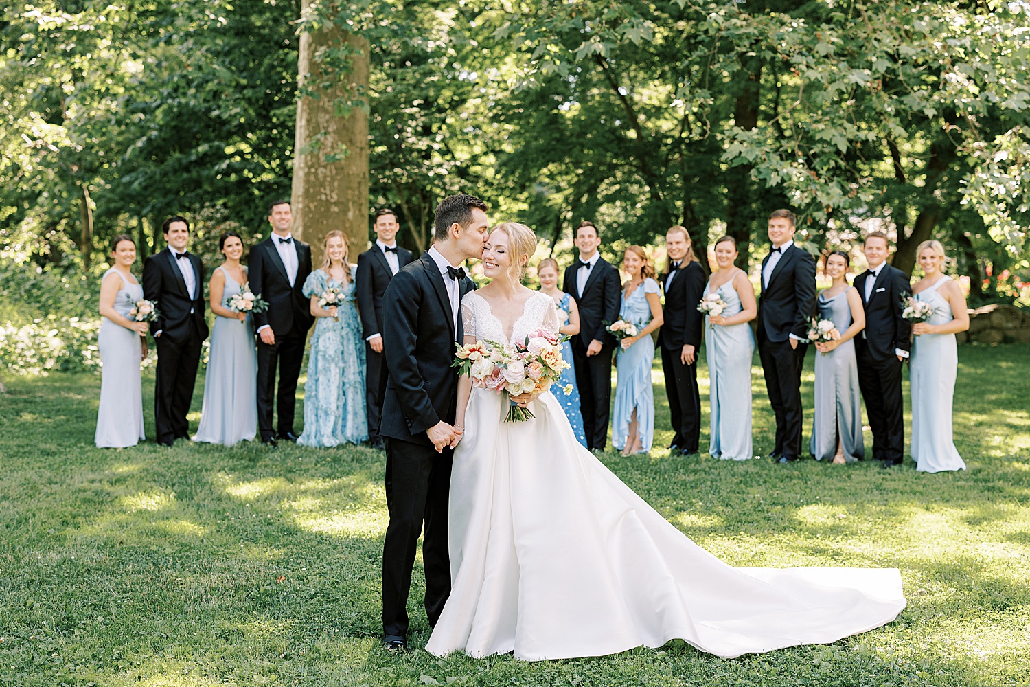 groom kisses bride's cheek in front of wedding party in black suits and blue dresses 