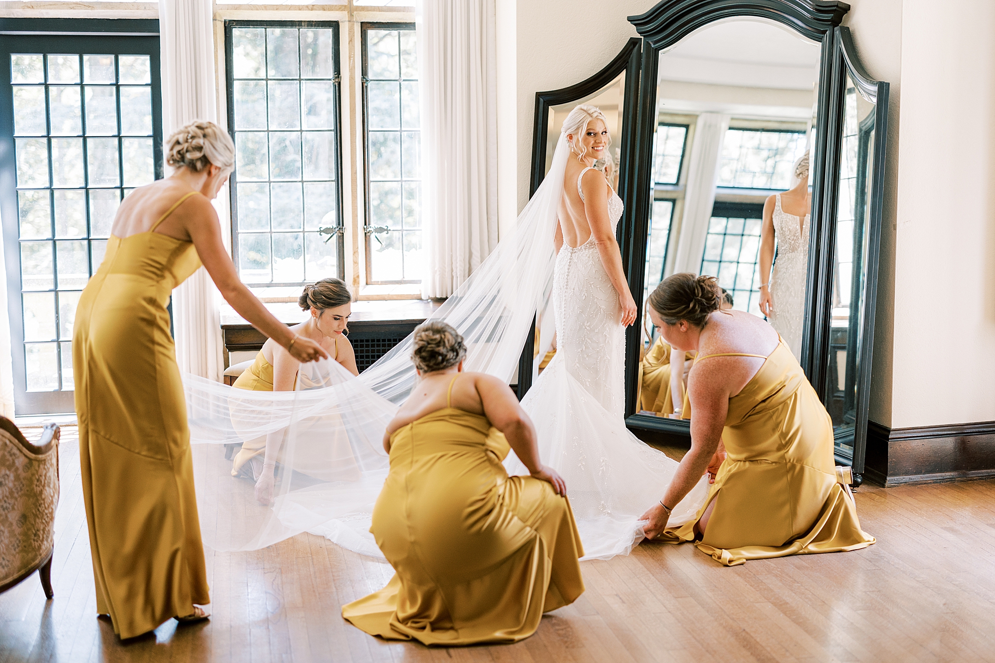 bridesmaids in gold help bride with veil and dress before wedding 