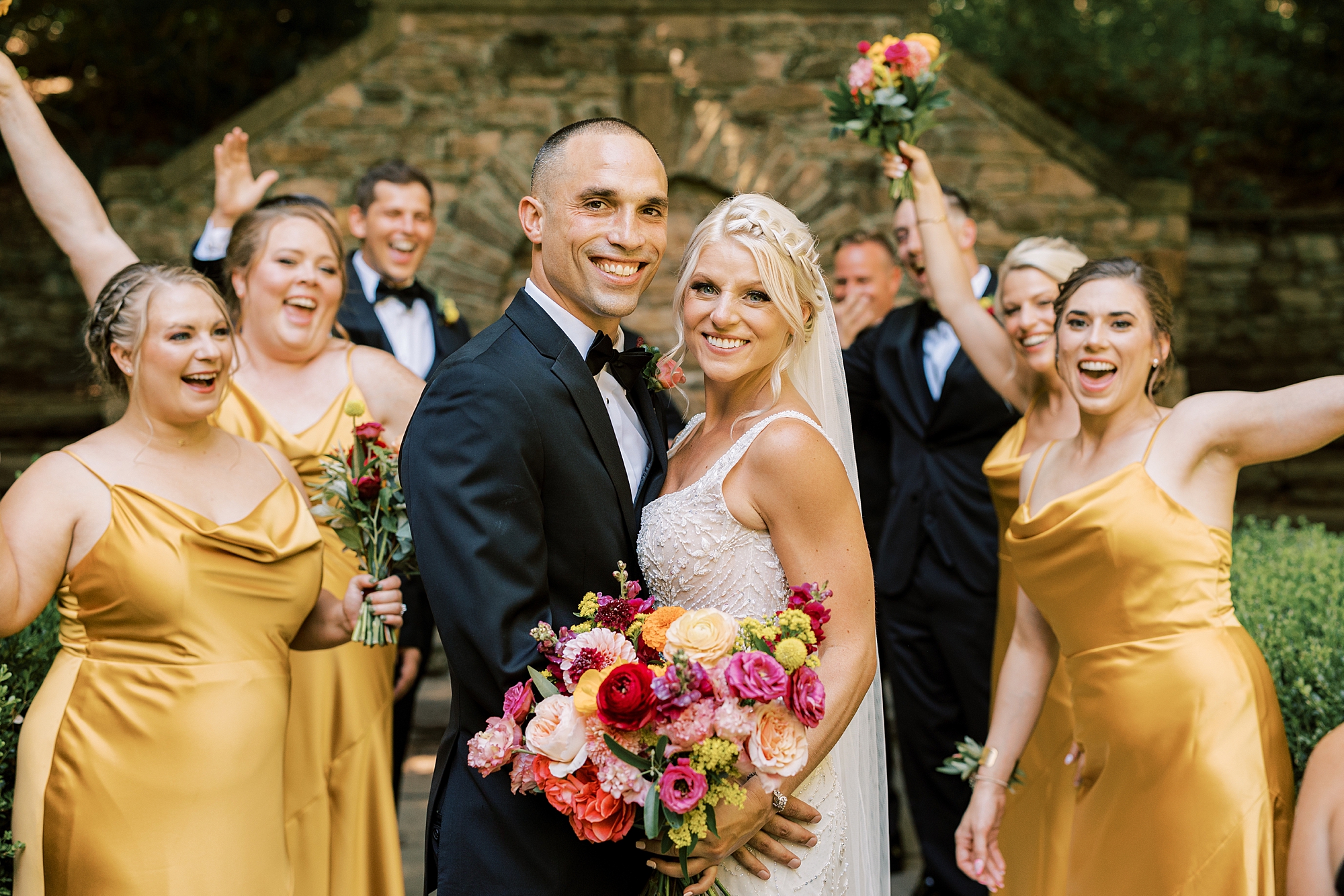 bride and groom hug with wedding party in gold and black attire behind them 