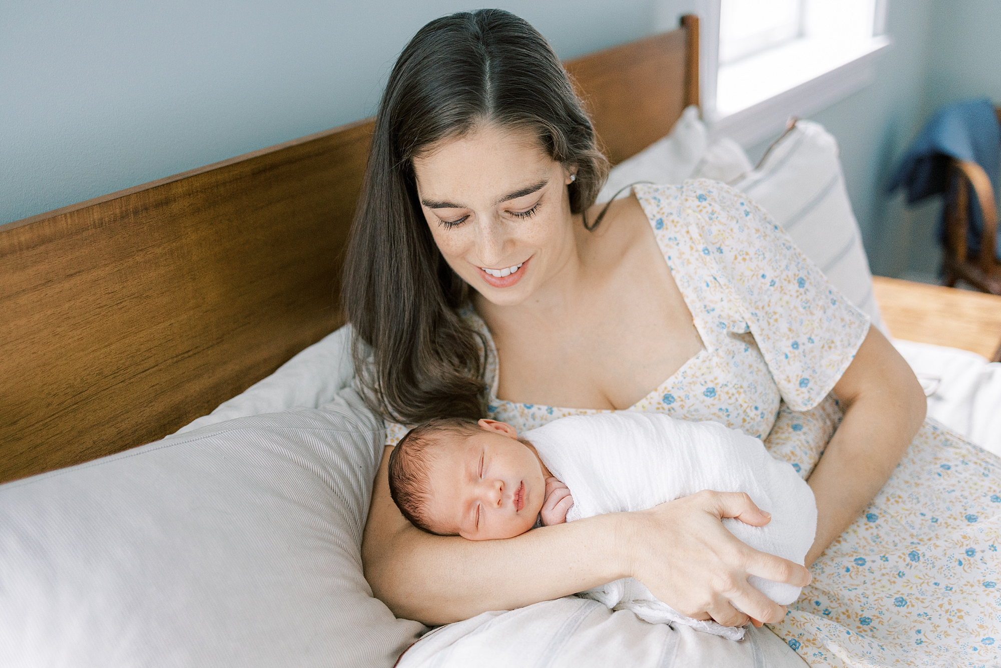 mom in white and blue dress holds newborn son in arms laying on bed