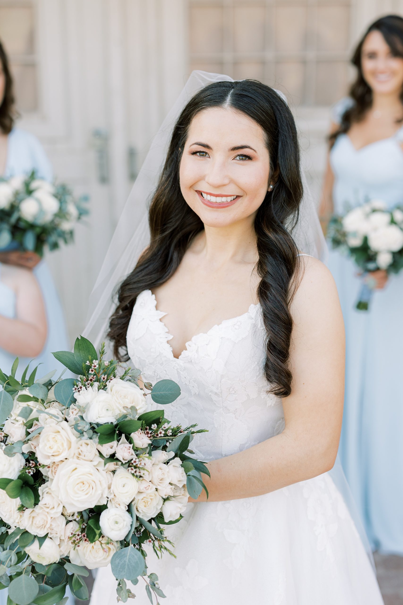 bride smiles holding bouquet of white flowers with bridesmaids in light blue gowns 
