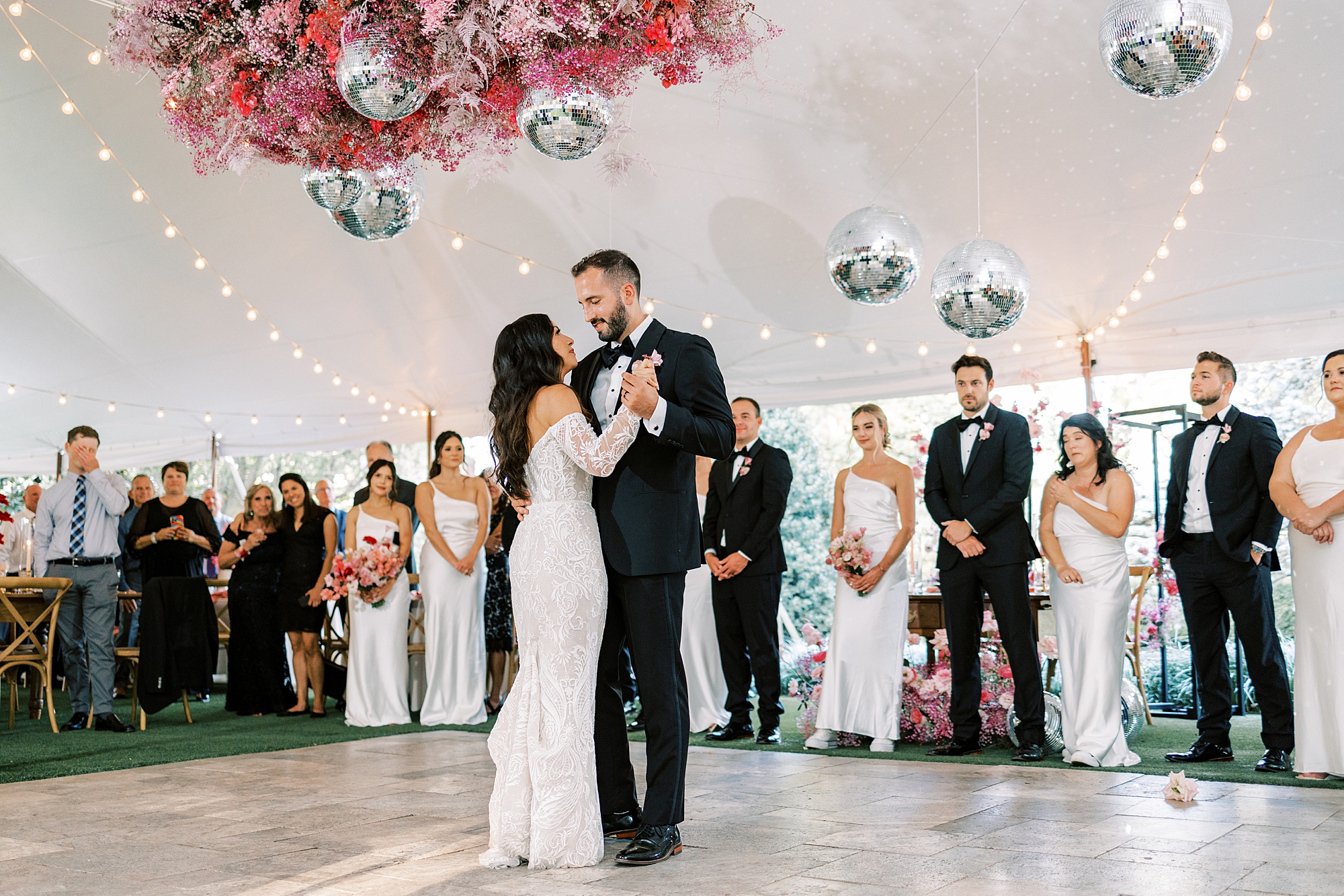 bride and groom have first dance on dance floor with monochromatic pink floral display and disco balls hanging from tent