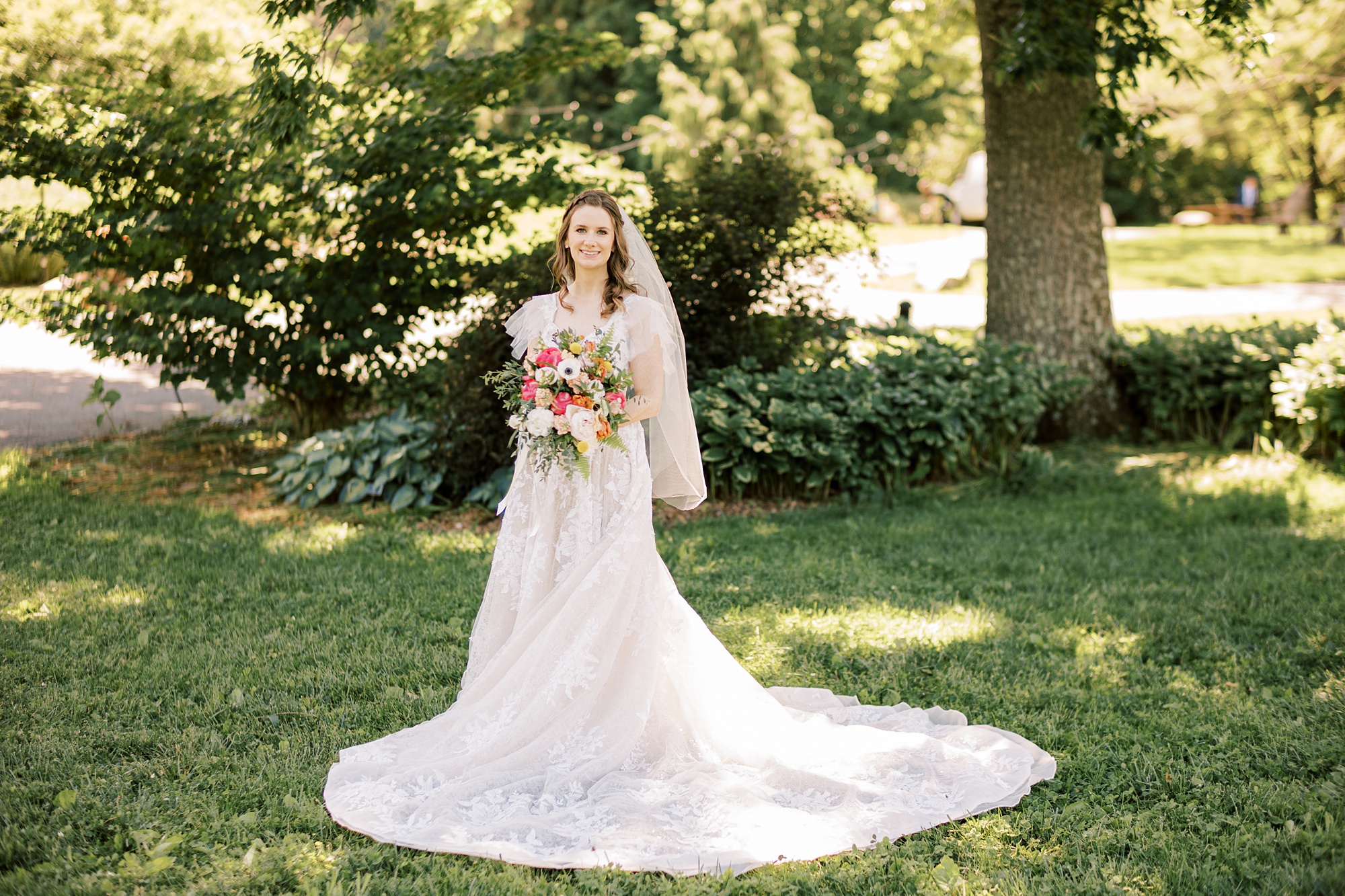 bride stands under tree holding bouquet with short veil around shoulders 