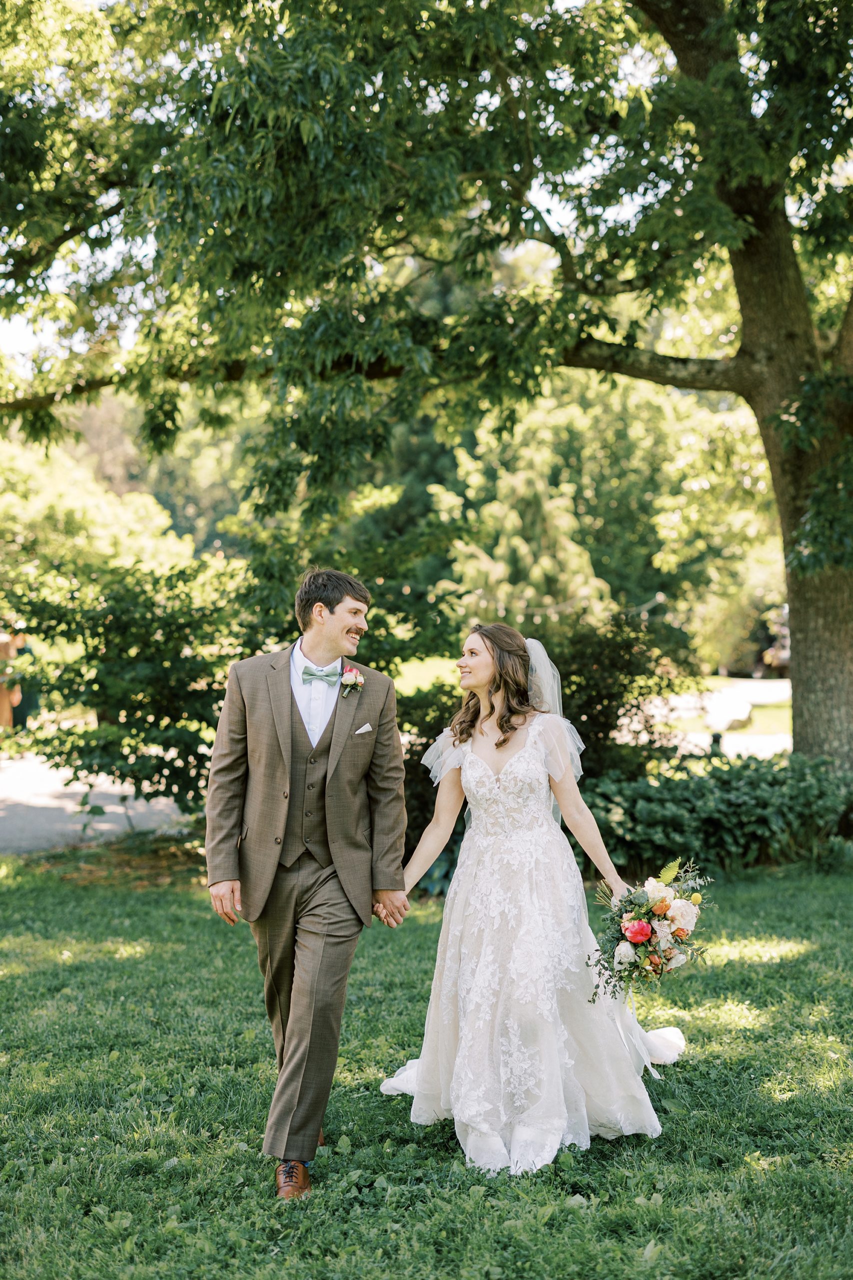 newlyweds hold hands walking through Tyler Arboretum during summer wedding day