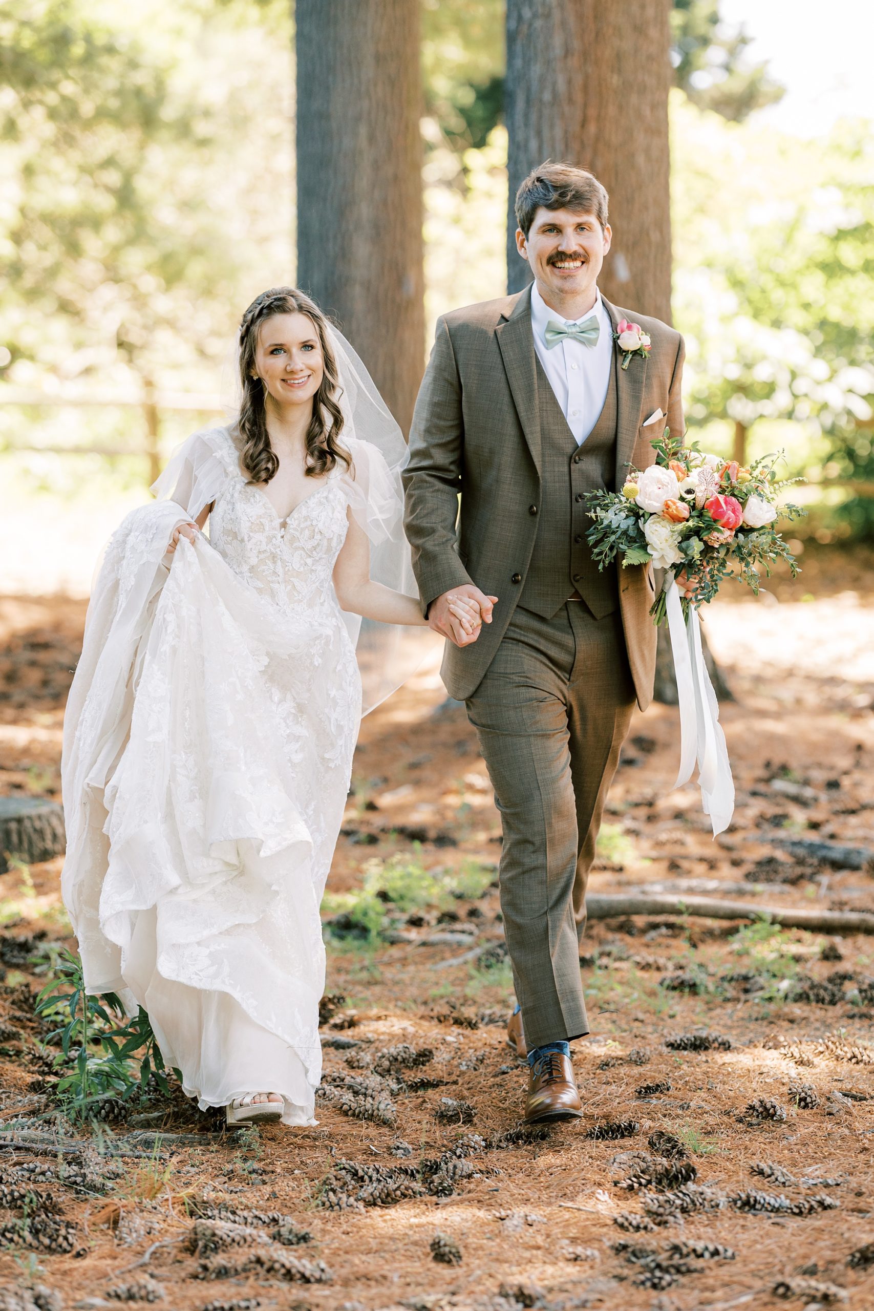 groom in brown suit carries bride's bouquet and helps her through the woods 