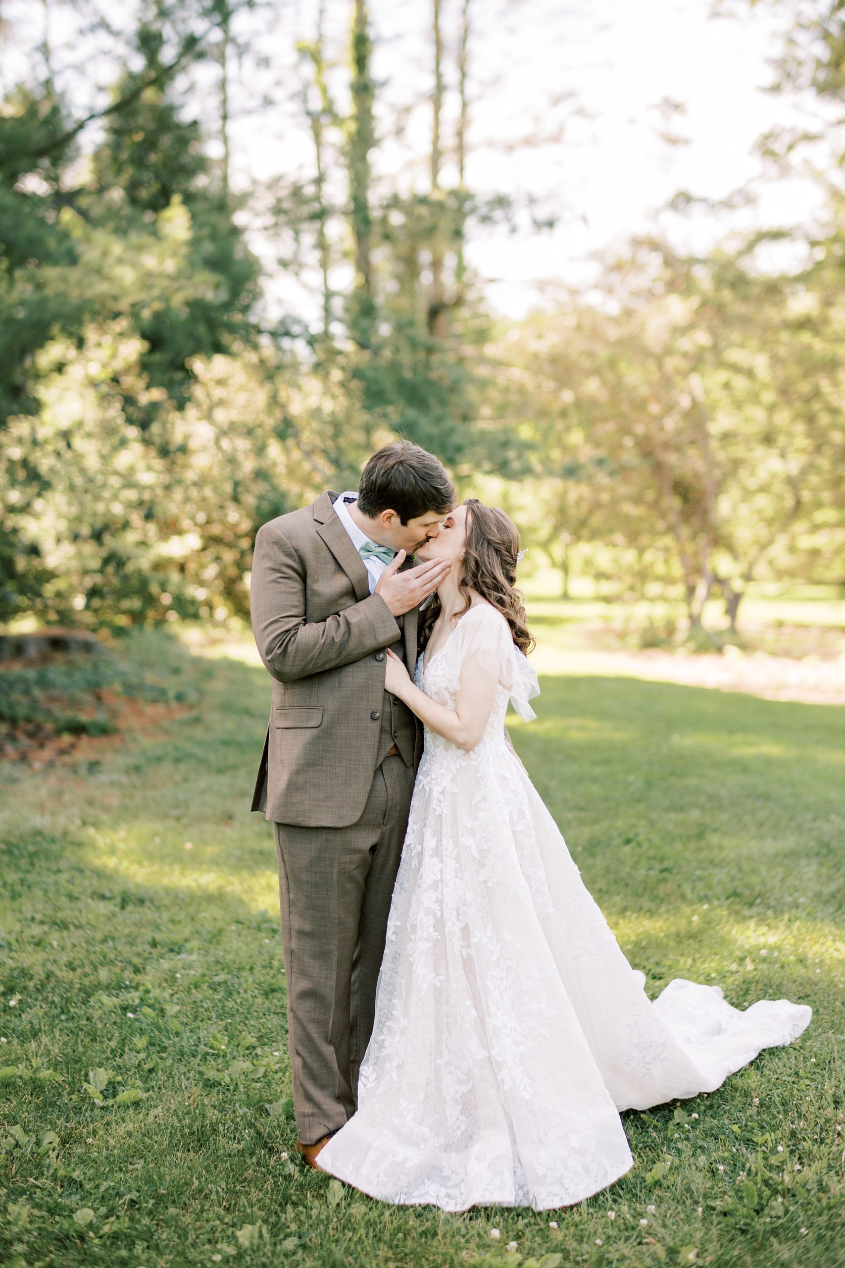 groom kisses bride holding her cheek during summer wedding portraits on lawn at Tyler Arboretum