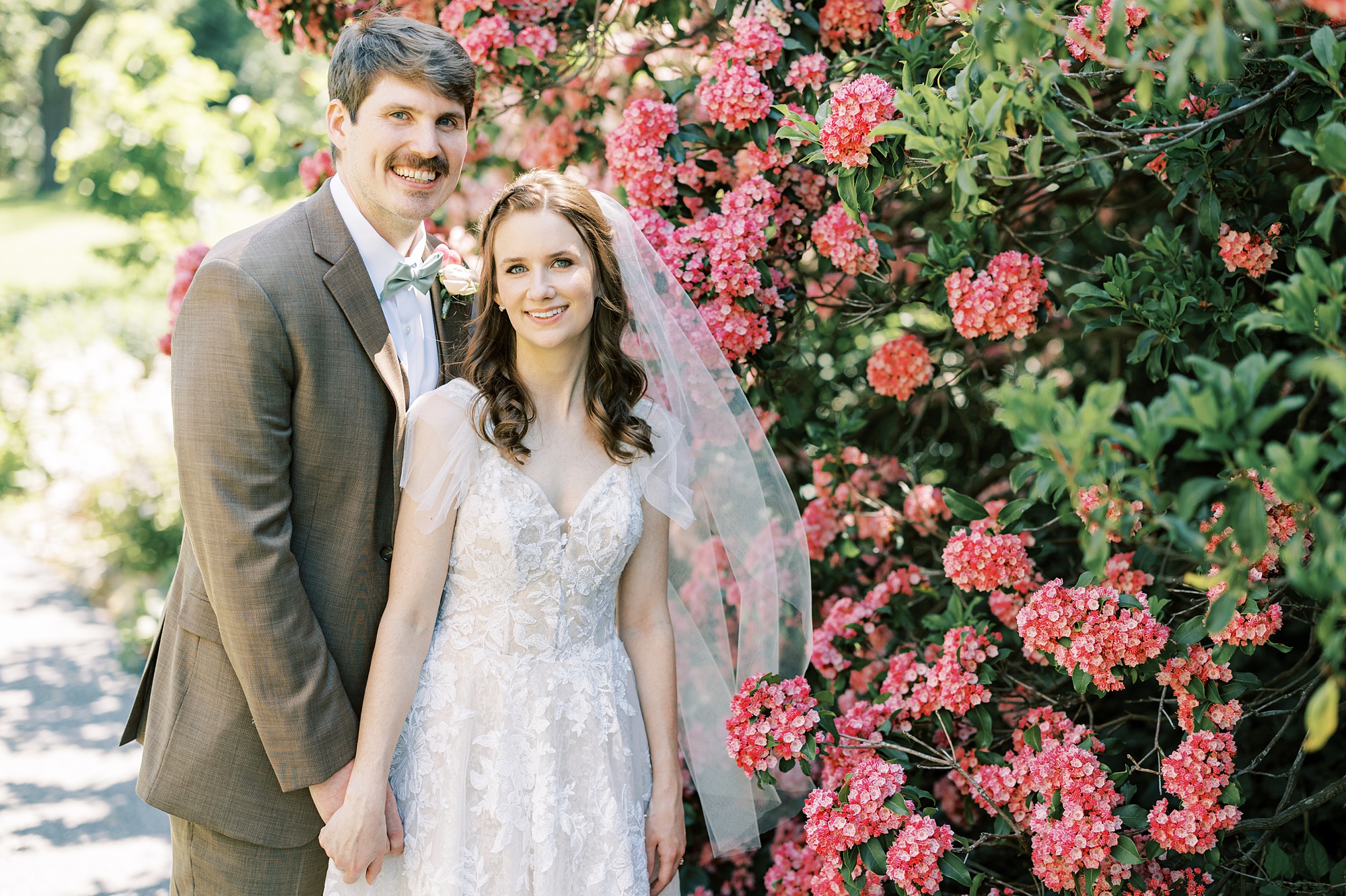 newlyweds hold hands and stand beside pink flower bush