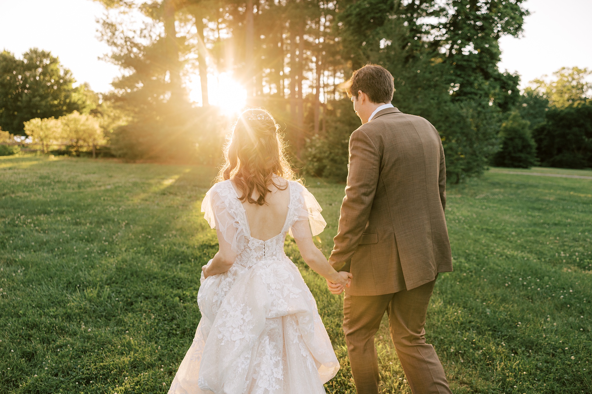 newlyweds hold hands walking through lawn at sunset 