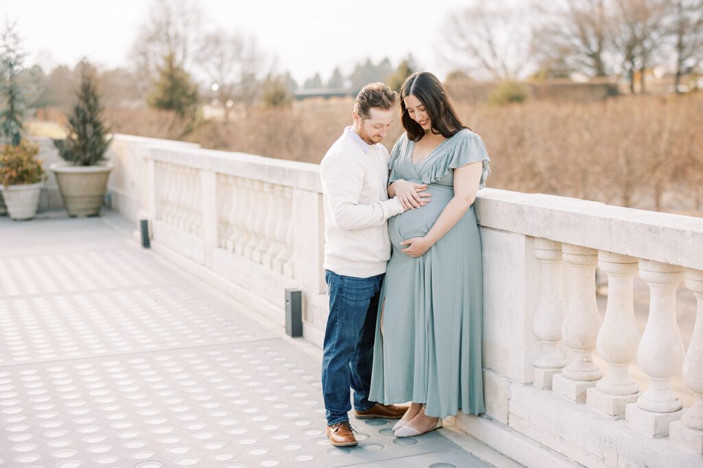 expecting couple looks at mom's baby bump on bridge at Longwood Gardens