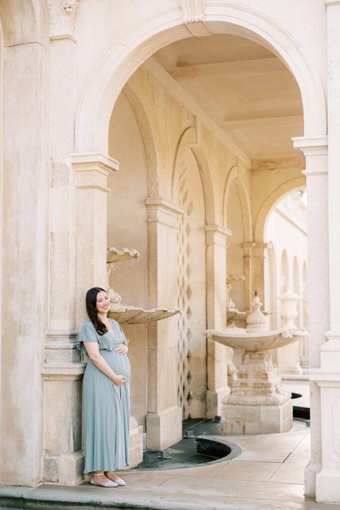 mother in sage green gown holds baby bump against white wall at Longwood Gardens