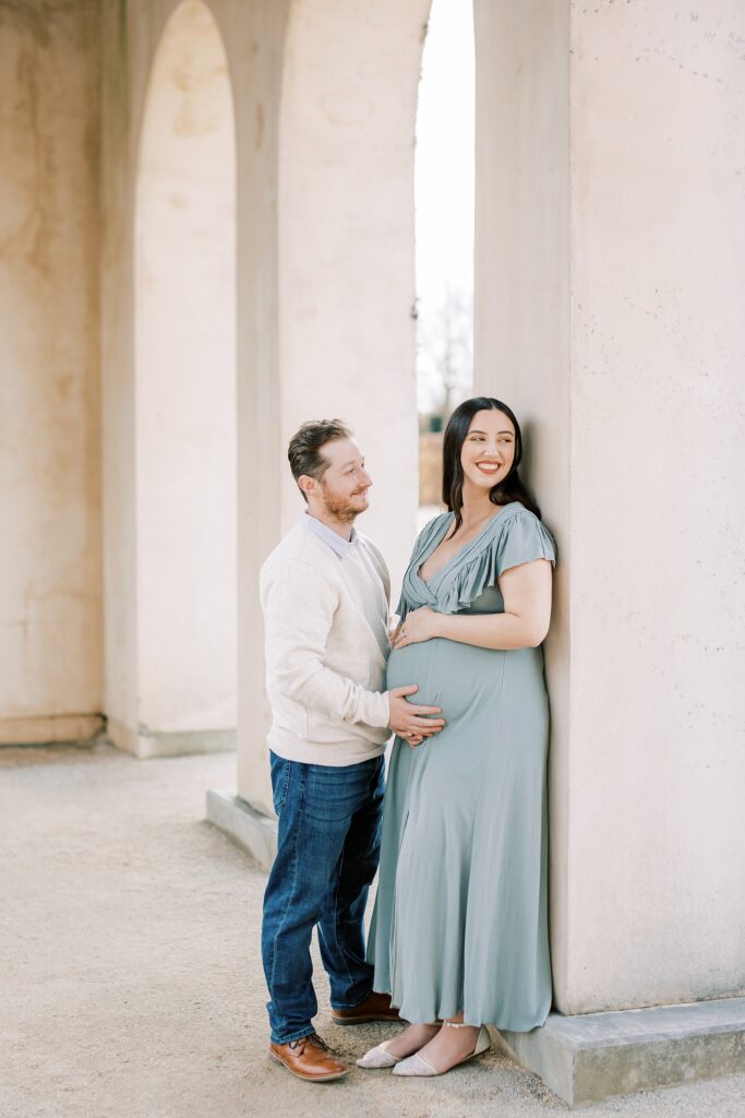 couple smiles and laughs leaning against pillar holding mom's baby bump