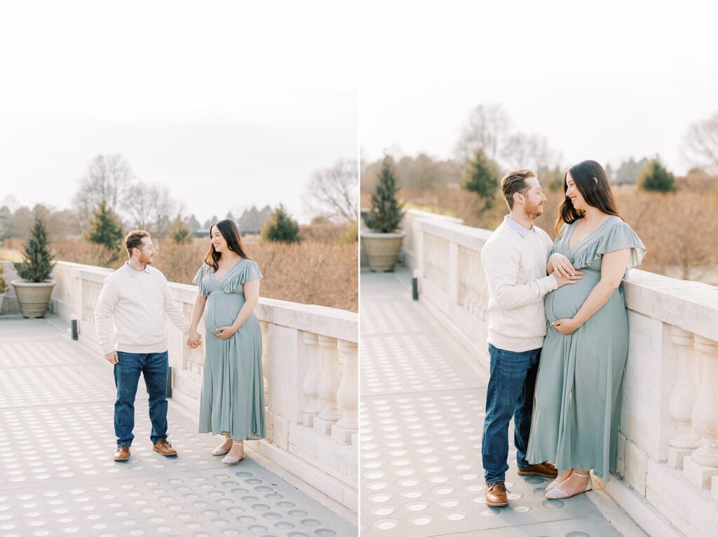 expecting parents walk on bridge in Longwood Gardens while mom holds stomach in sage green gown