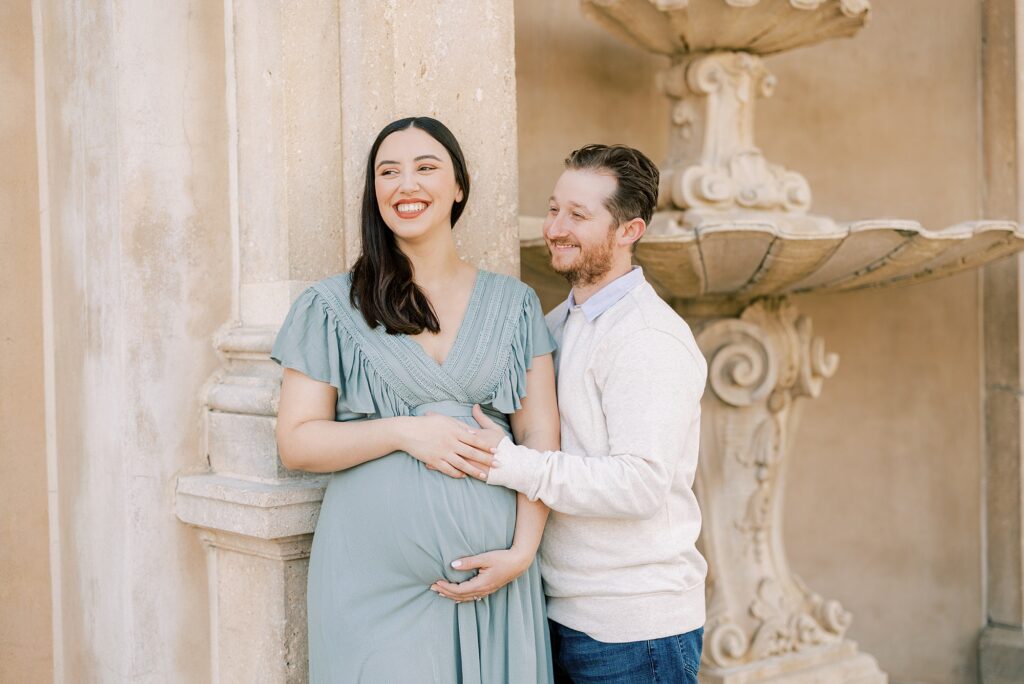 man laughs with wife in sage green gown holding her baby bump near fountain