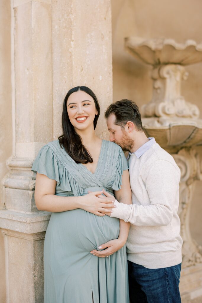 man leans into woman's shoulder in sage green gown laughing while holding her baby bump