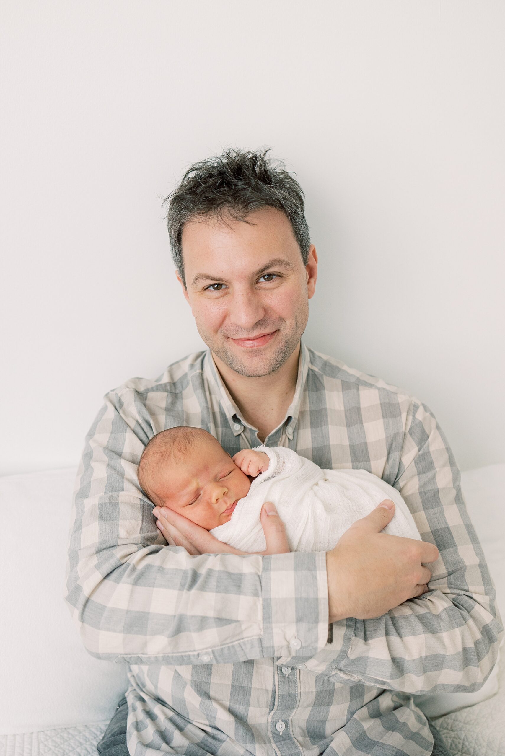 man in green and white plaid shirt holds son in white wrap on his chest 
