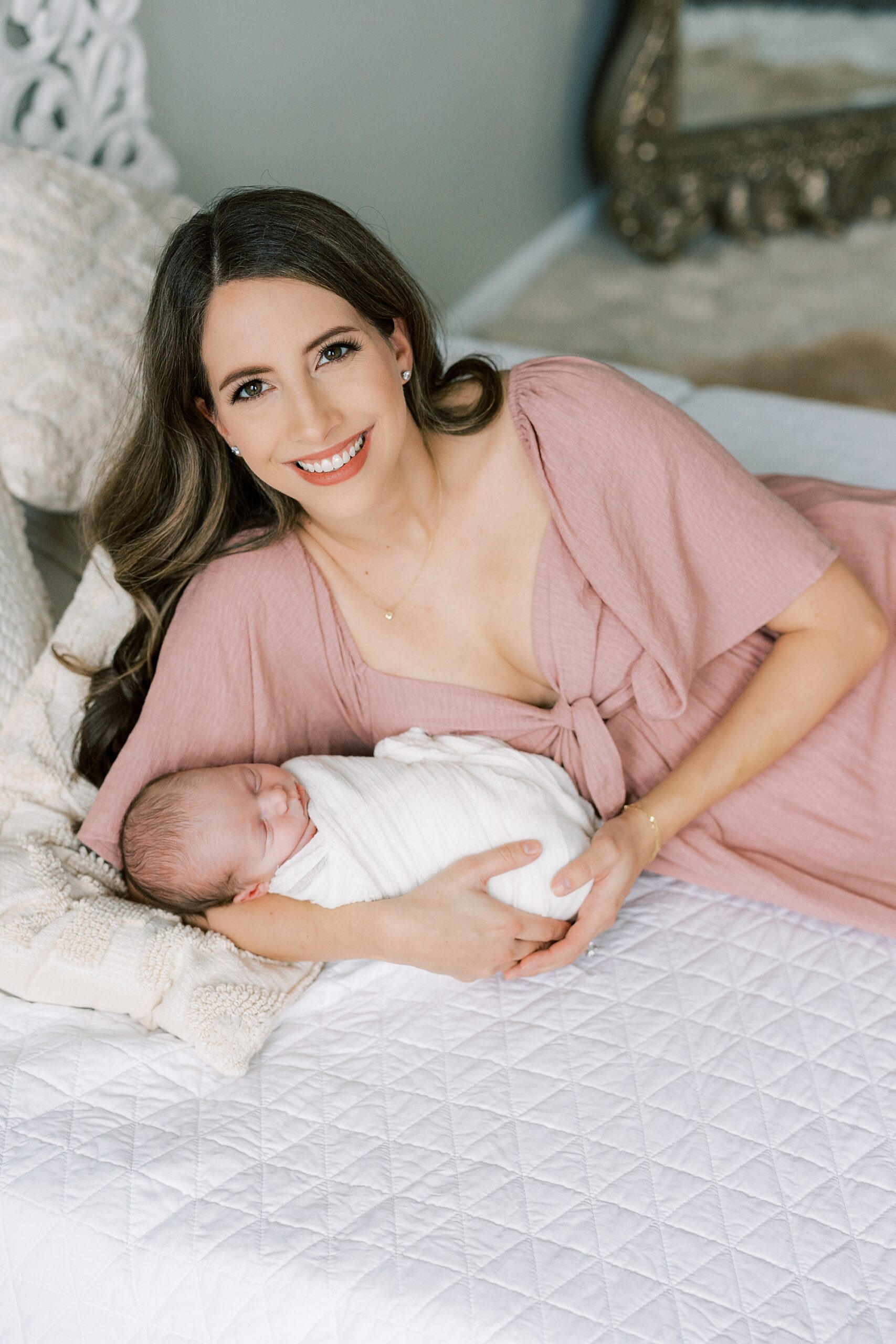 mom in pink dress lays on bed with newborn baby girl 
