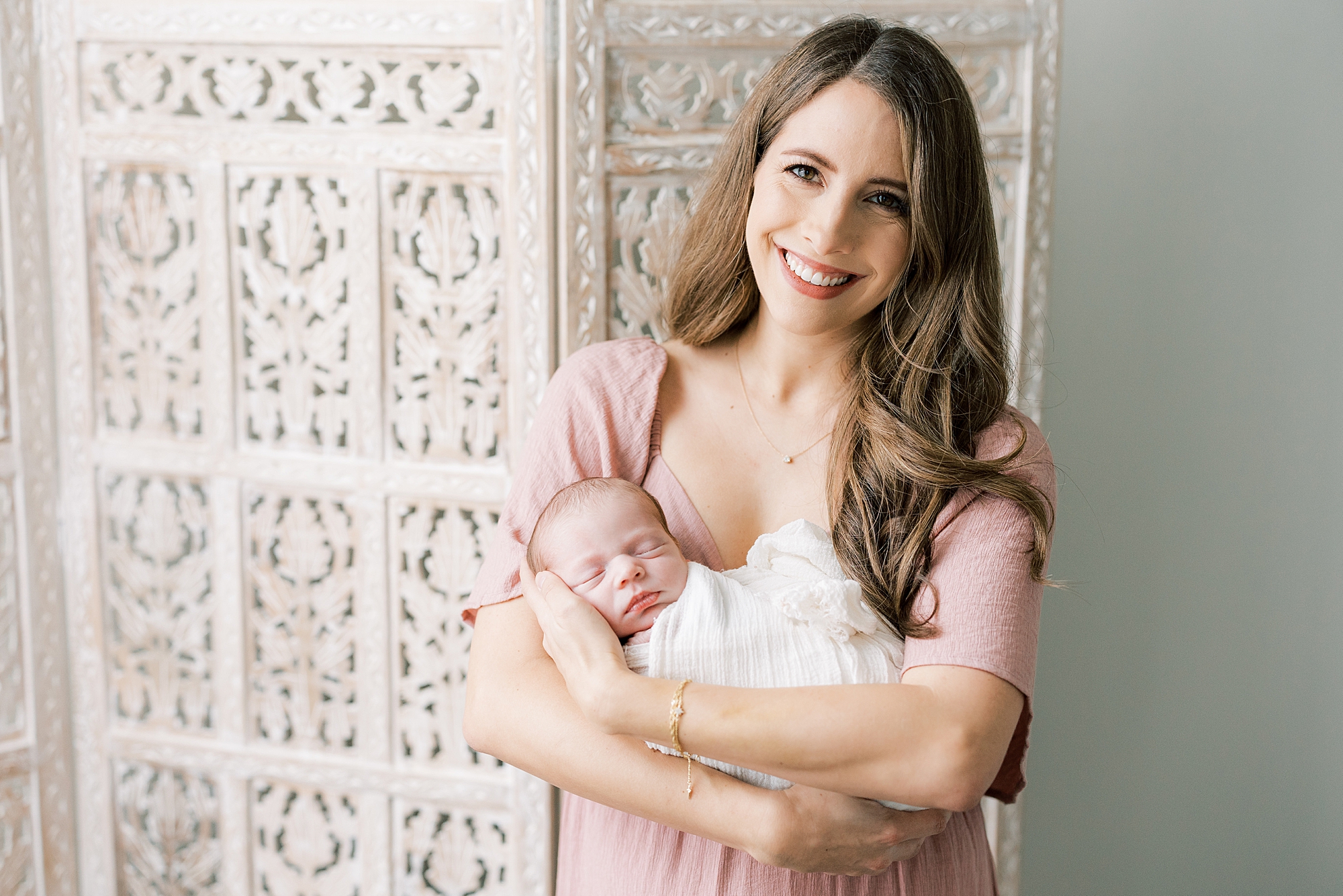 mother in pink dress holds baby girl next to wall divider 