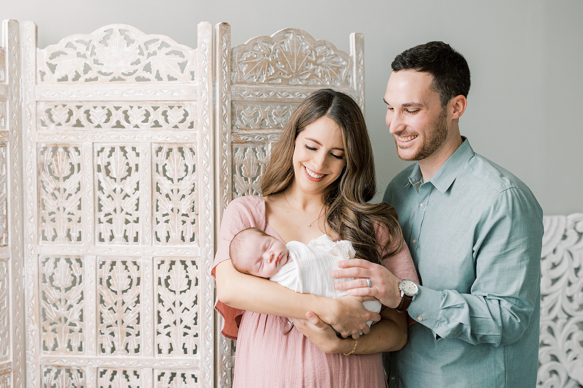 new parents hold baby girl in white wrap standing by room divider 