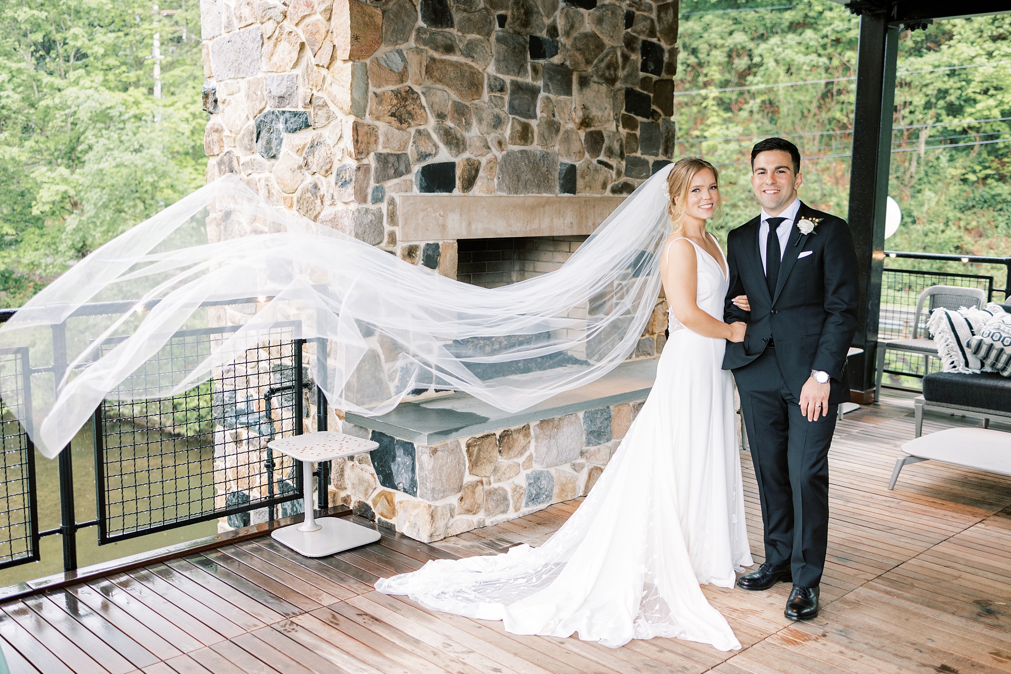 bride and groom pose by stone fireplace while bride's veil floats behind them