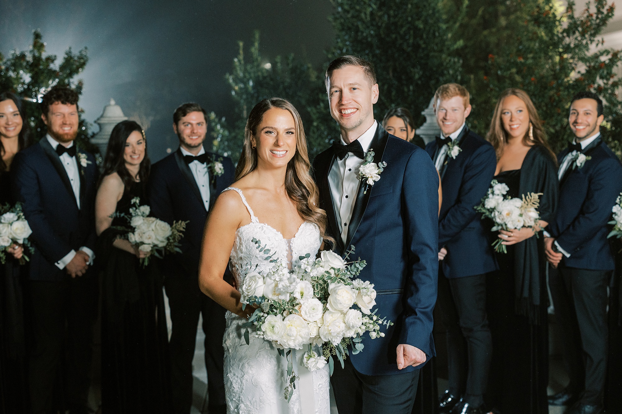 newlyweds stand together in front of wedding party in black dresses and blue suits