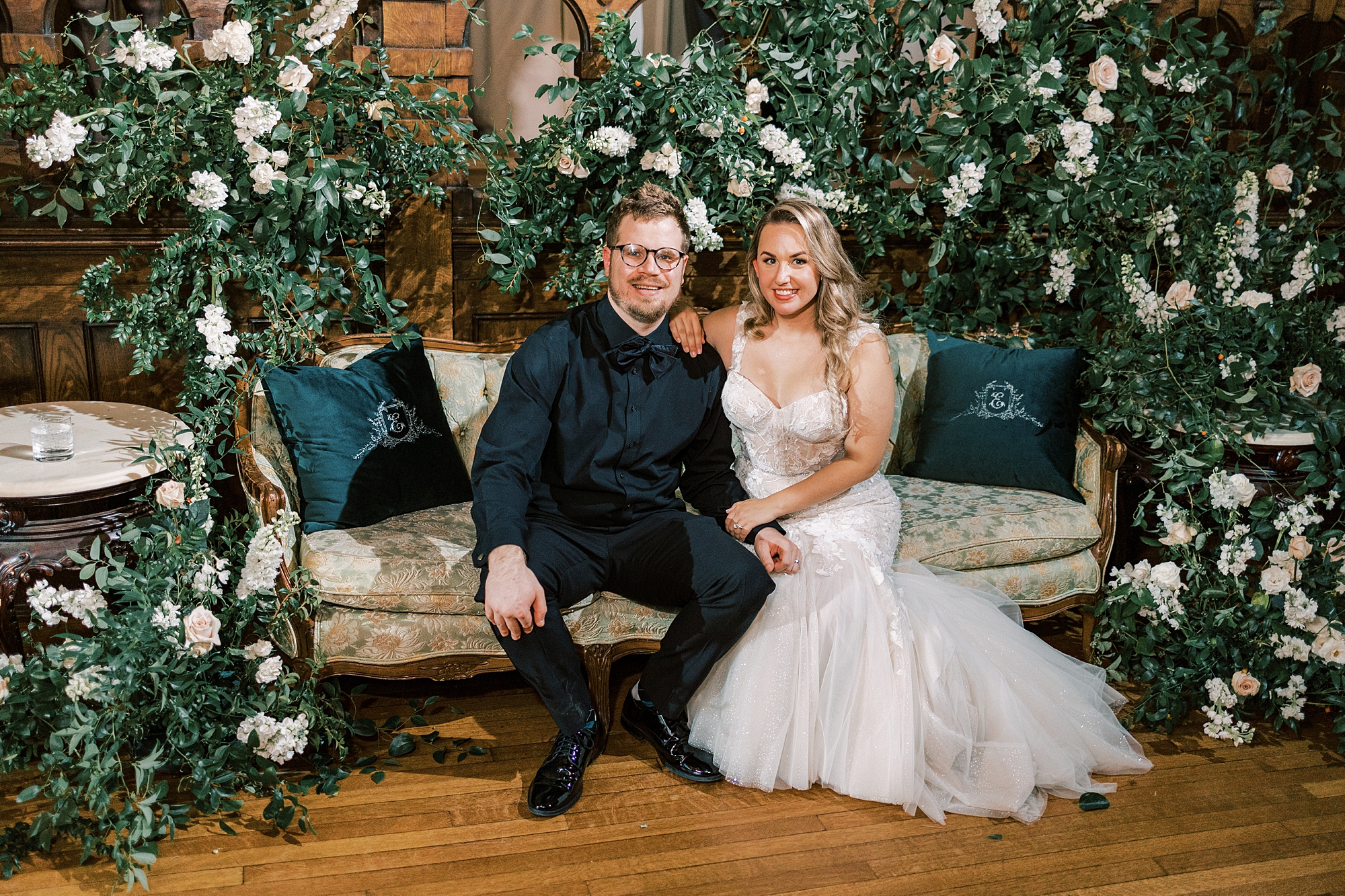 newlyweds sit on sofa with floral arrangement above them 