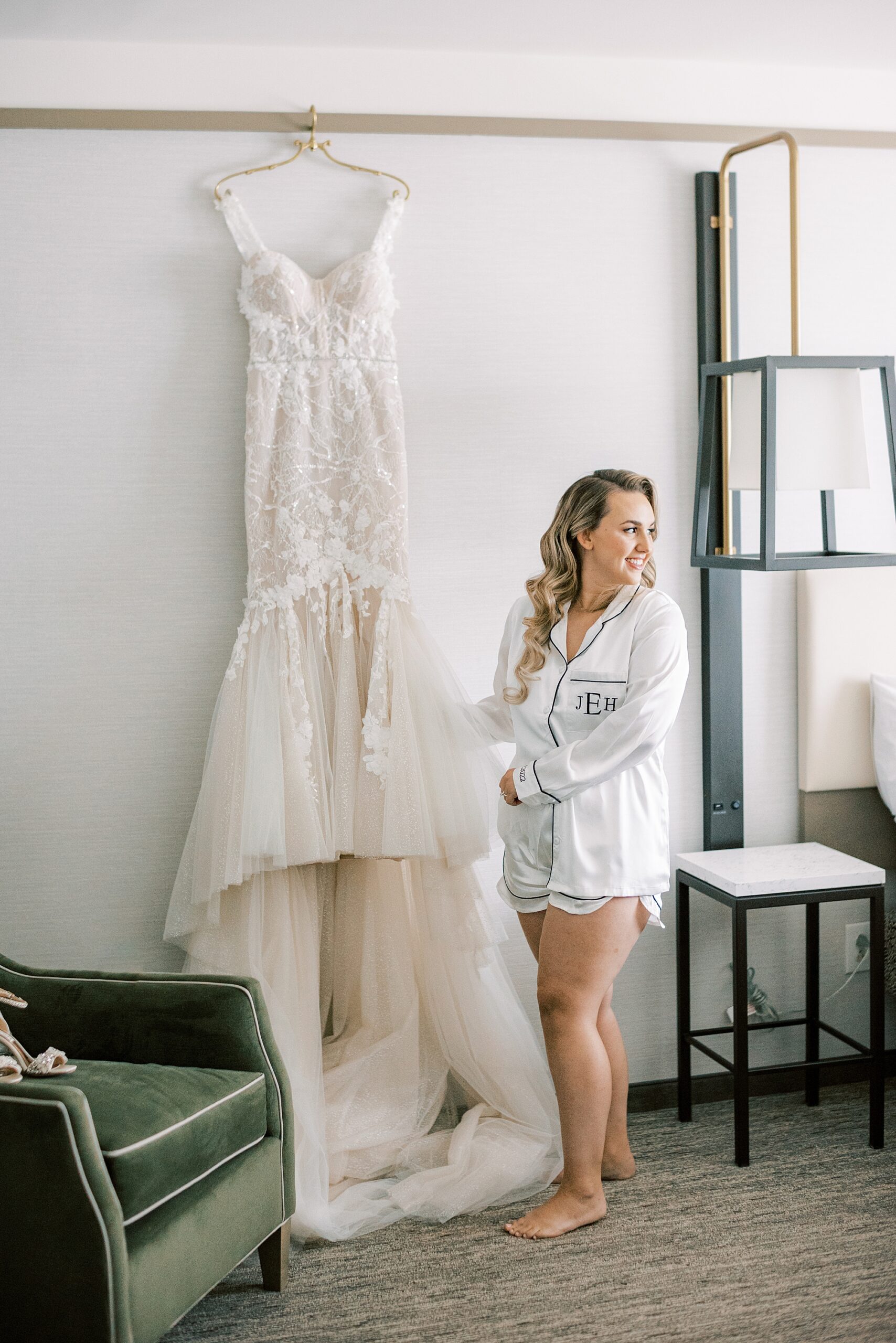 bride poses next to wedding gown hanging on white wall 