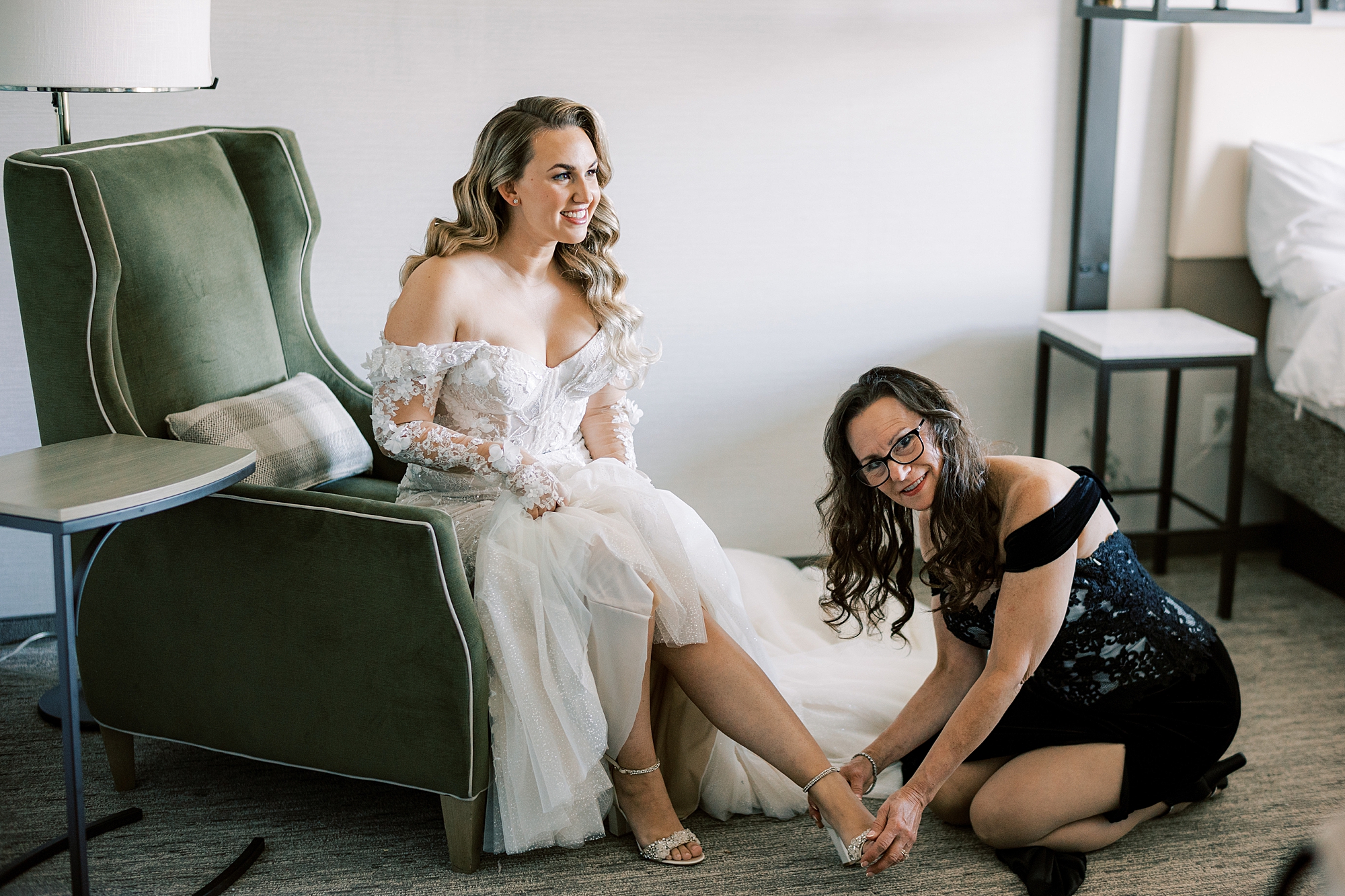 bridesmaid in black skirt kneels to help bride with shoes 