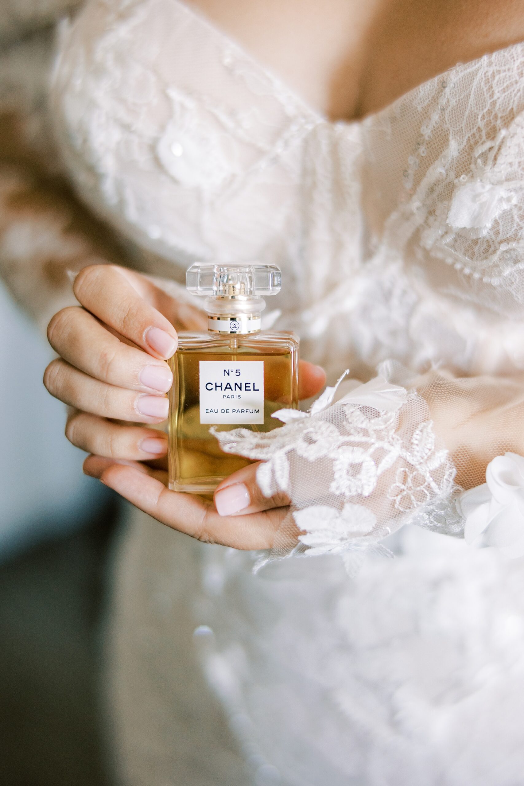 bride holds bottle of perfume in front of wedding gown with lace overlay 