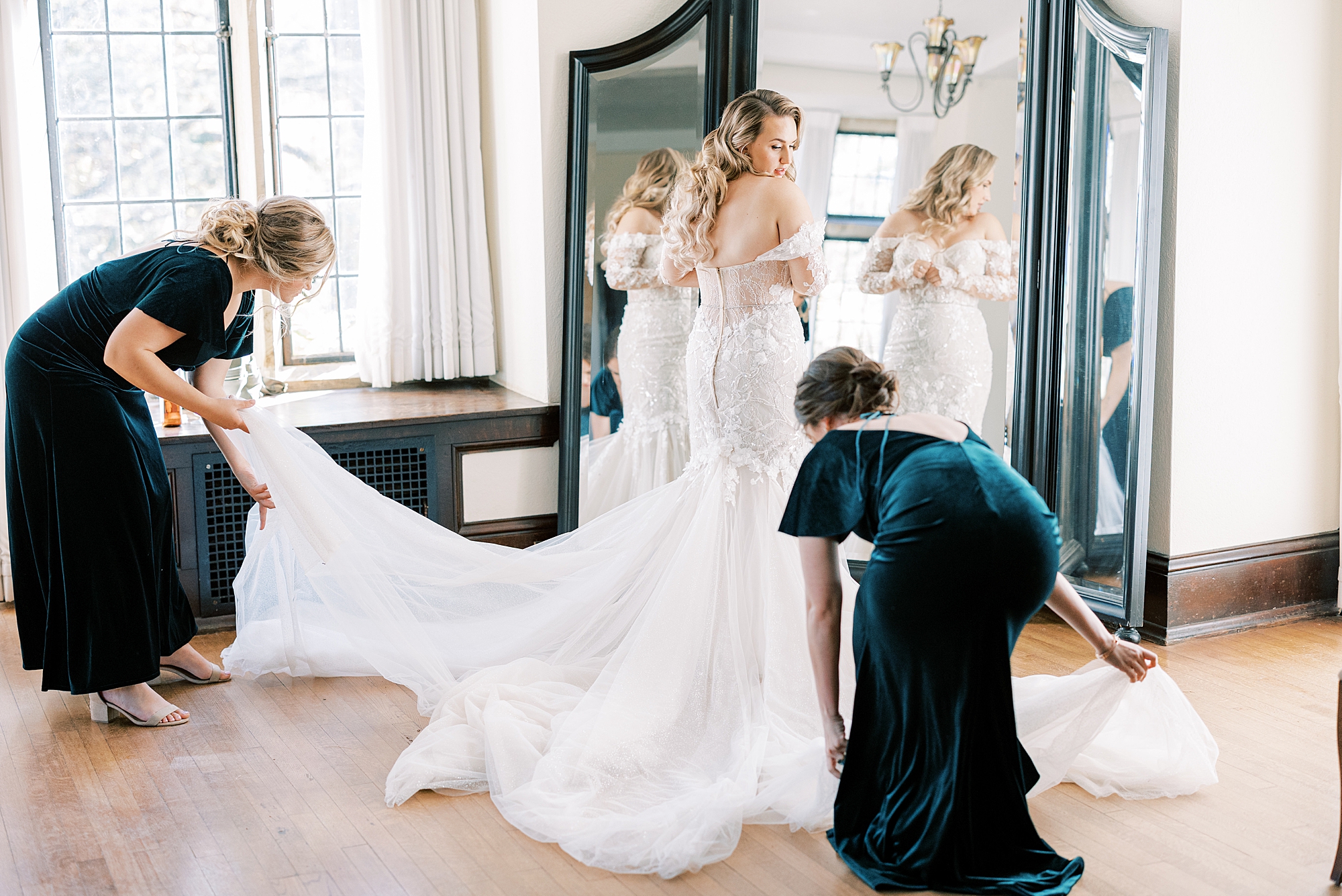 mother and bridesmaid adjust bride's veil and skirt in front of mirror 