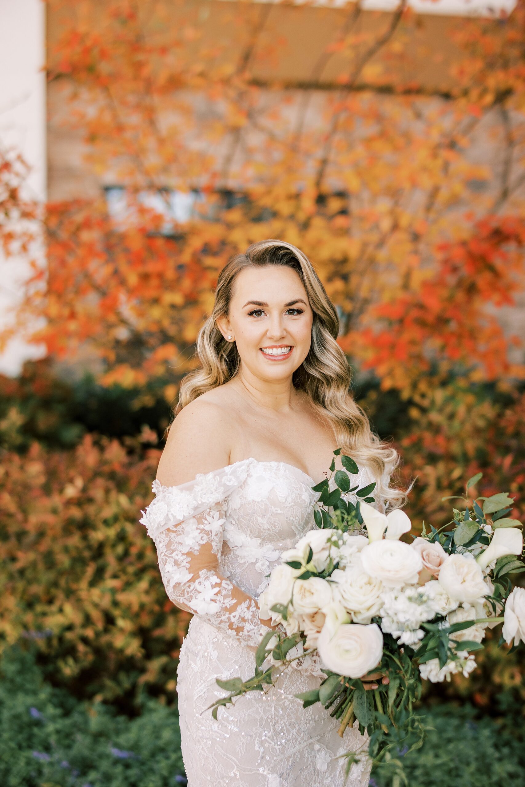bride stands holding pink and white rose bouquet by orange laves on trees 