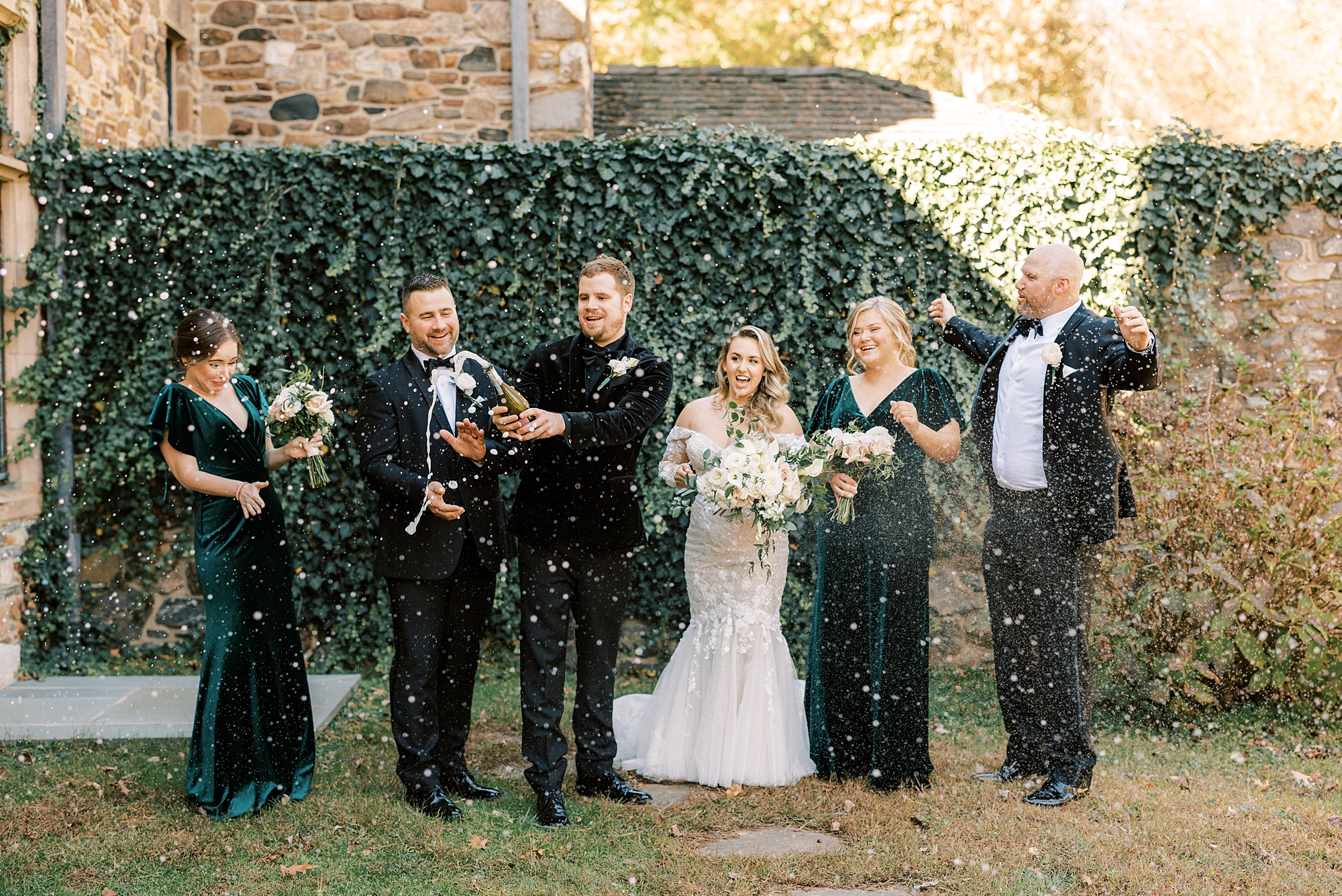 bride and groom pop Champagne with wedding party along greenery wall 