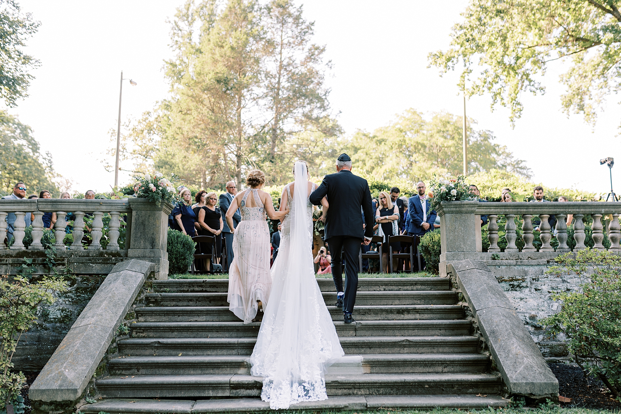 bride walks up steps for Curtis Arboretum wedding ceremony on lawn with parents 