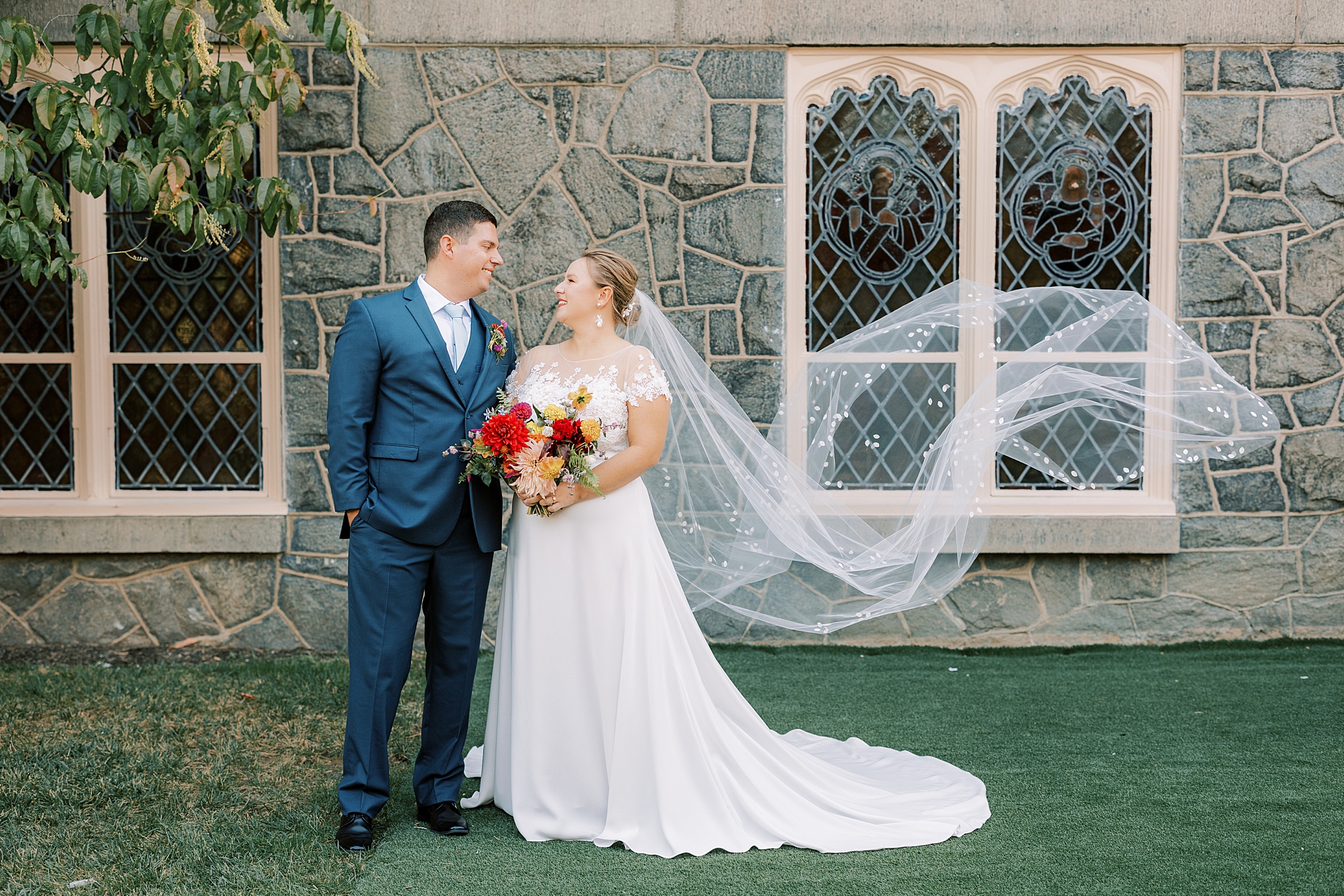 bride and groom smile at each other by stone windows at St. Ann’s Roman Catholic Church